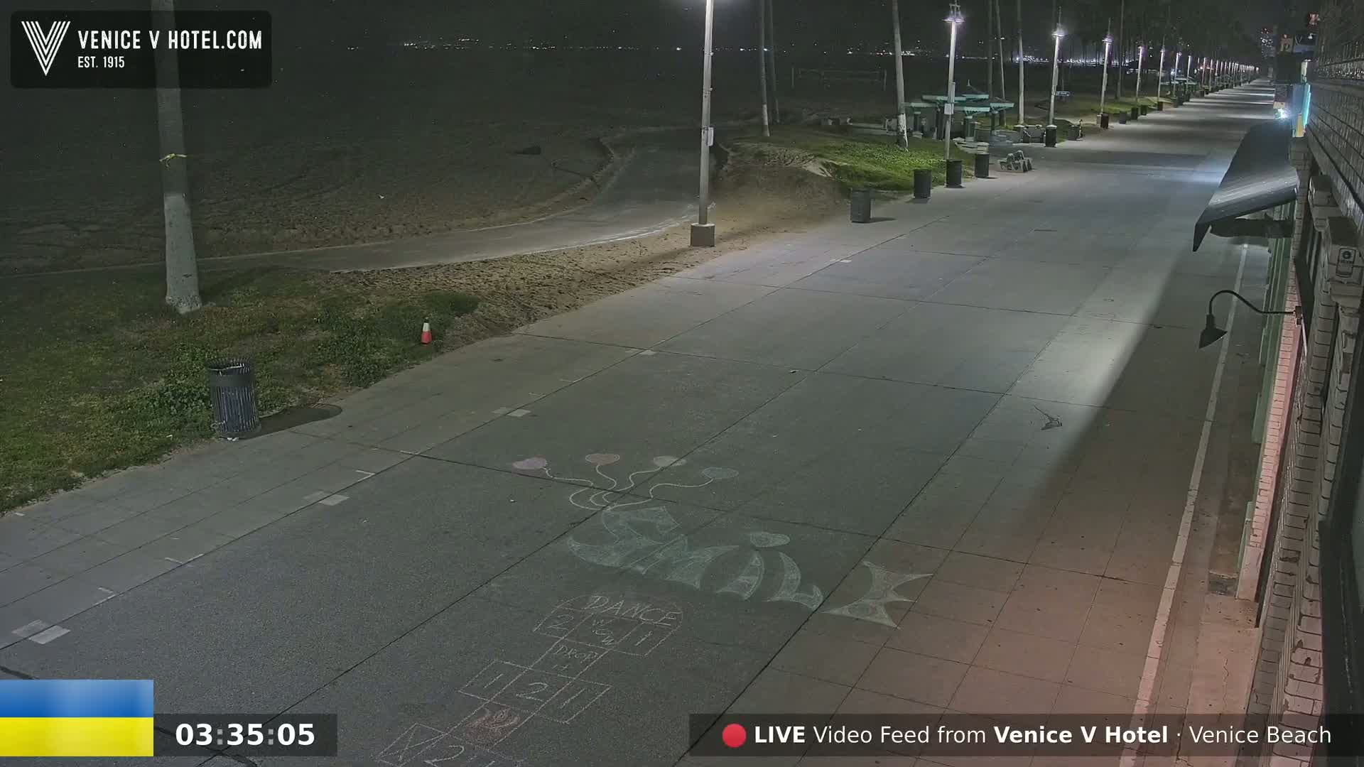 A nighttime view of a mostly empty boardwalk next to a sandy beach, illuminated by streetlights, with children's chalk drawings on the pavement.
