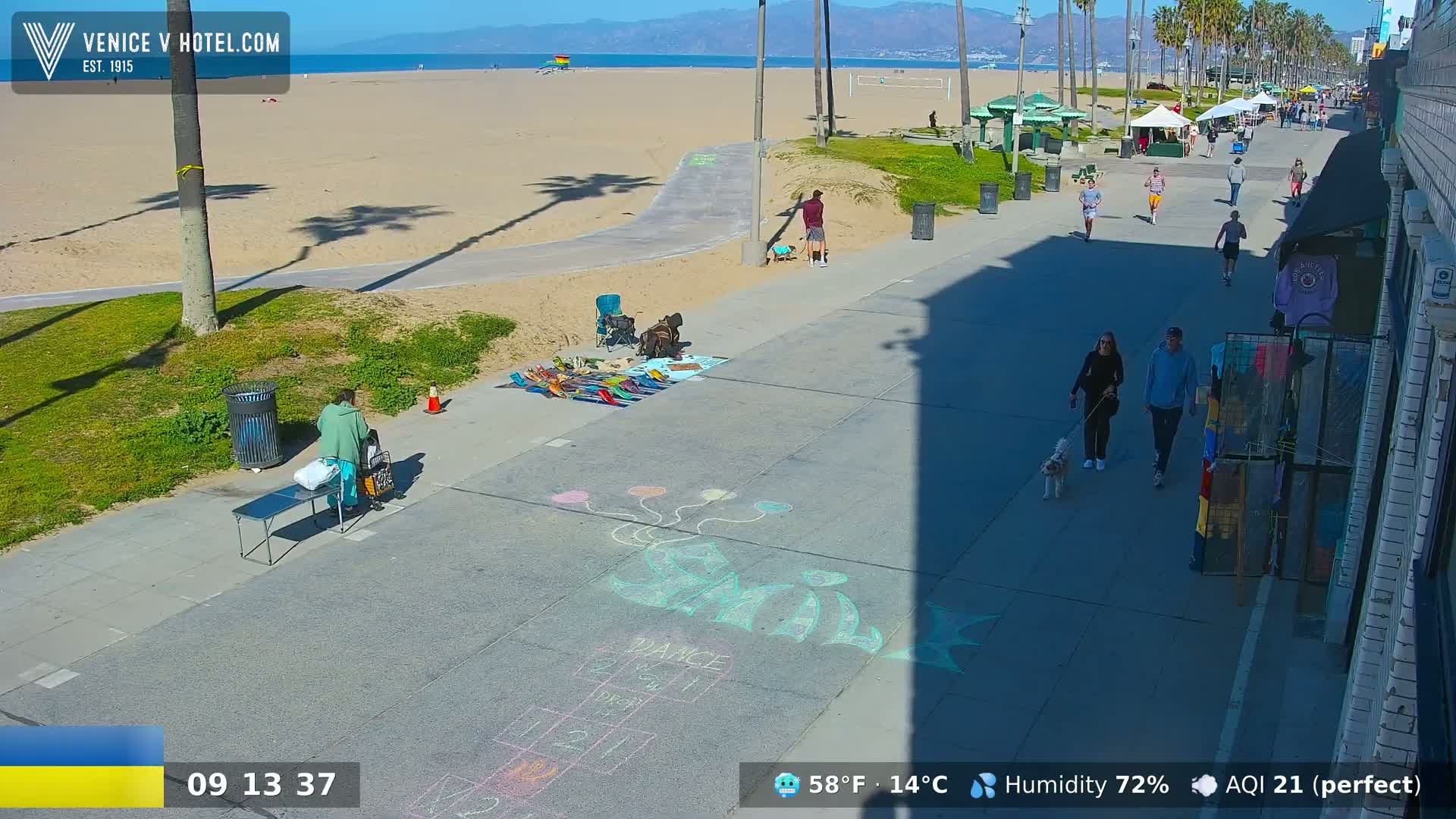 A sunny beach boardwalk scene shows several people walking, a vendor selling goods, and chalk drawings on the pavement.