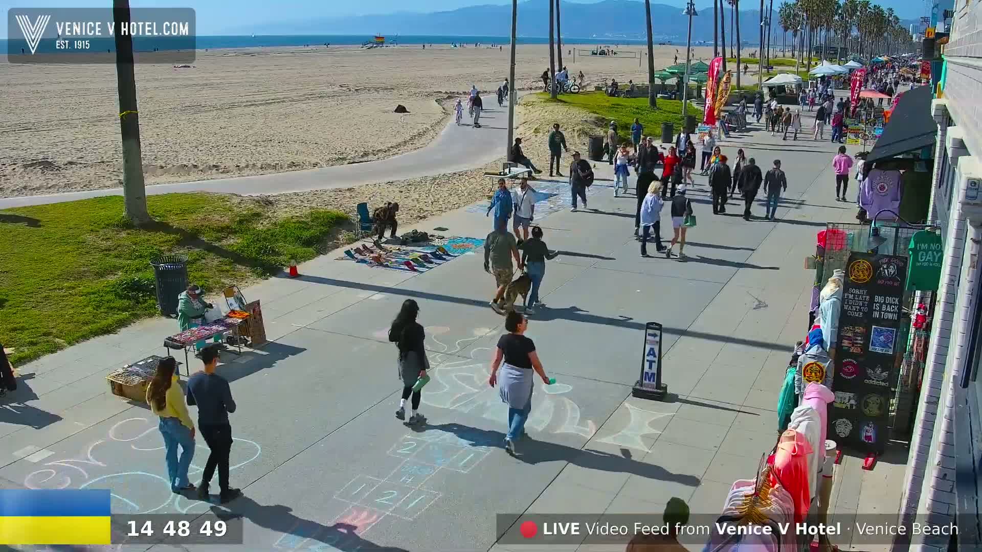A sunny beach boardwalk scene shows many people walking past vendors and chalk drawings on the pavement, with a sandy beach and ocean in the background.