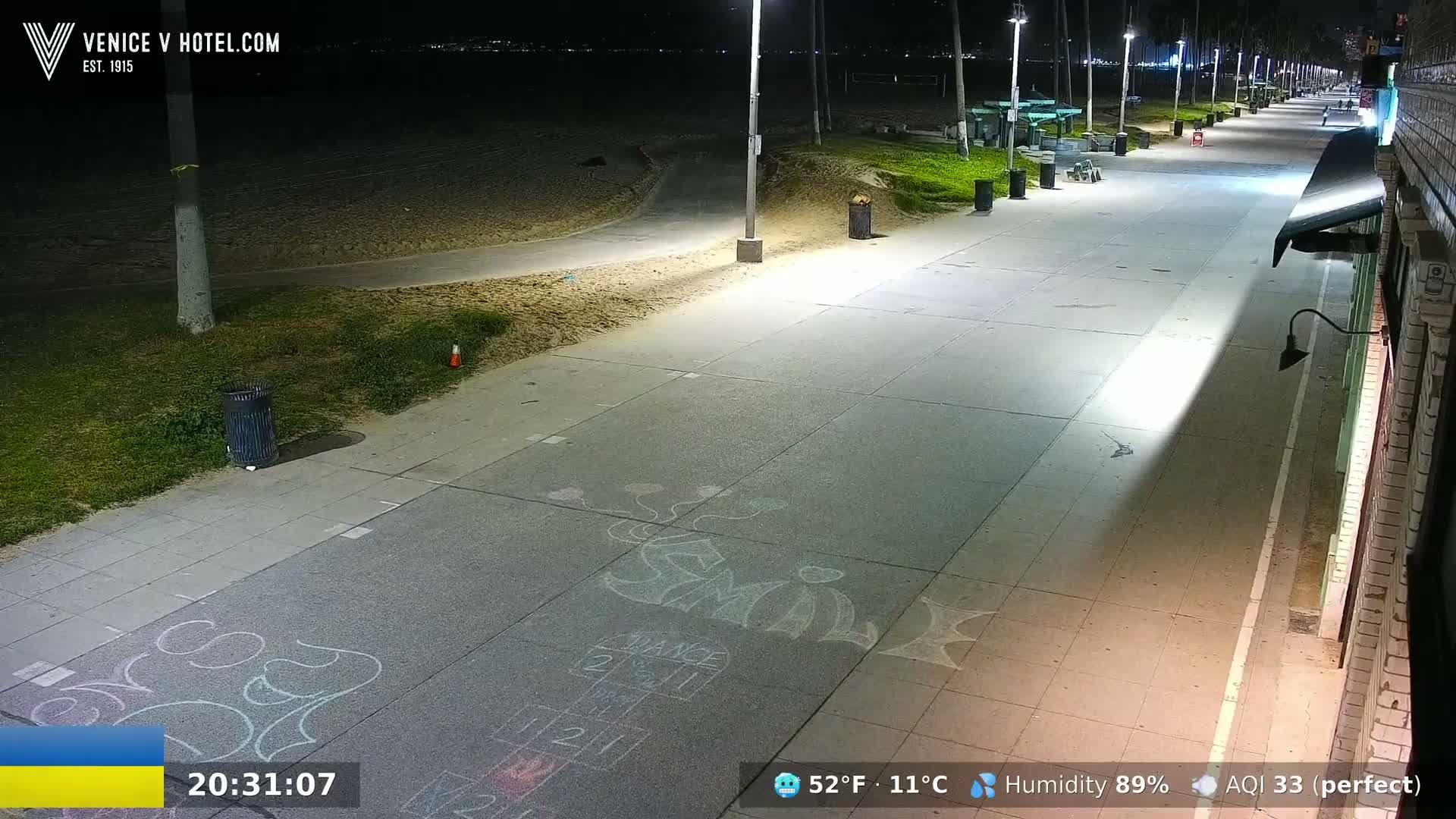 A nighttime view of a paved beachfront walkway with chalk drawings on the ground, illuminated by streetlights, next to a sandy beach.