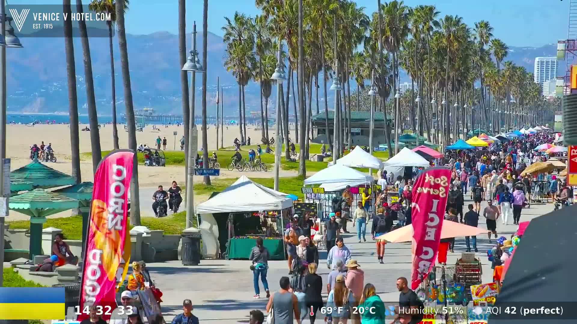 A sunny day at a beach boardwalk with many people walking among numerous vendors and palm trees.