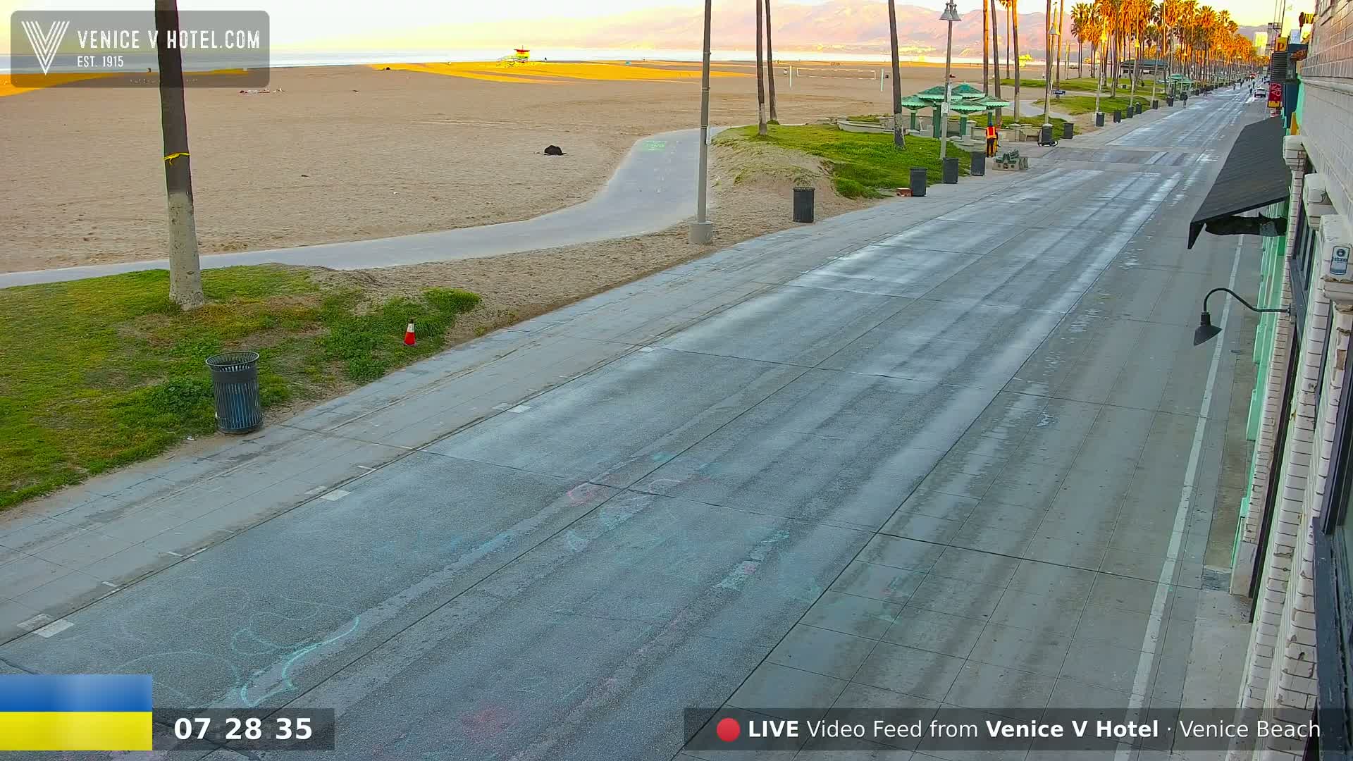 A mostly empty paved beachfront road stretches towards a sandy beach with a lifeguard stand under a clear, bright sky.