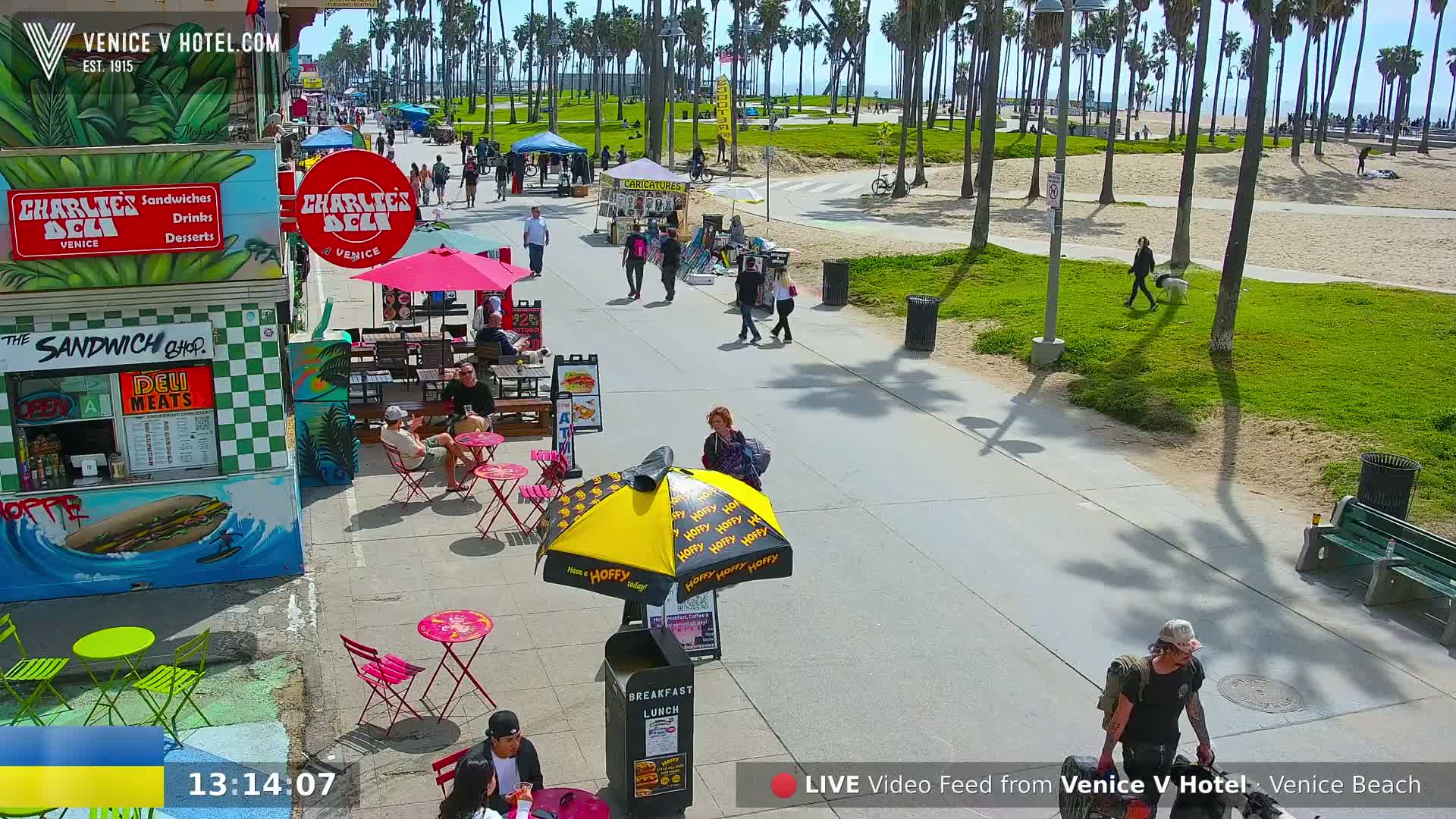 A sunny boardwalk scene shows people walking and sitting at outdoor cafes beneath palm trees, near a beach.