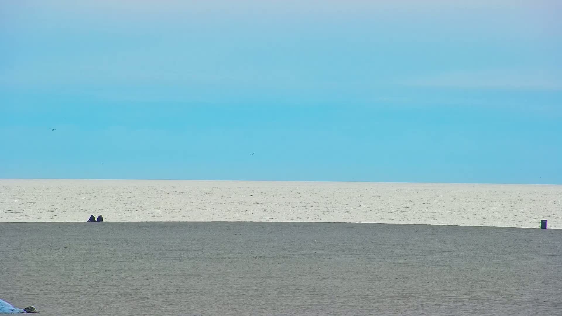 Two figures sit on a grey beach beneath a pale blue sky and a calm, light-grey sea.