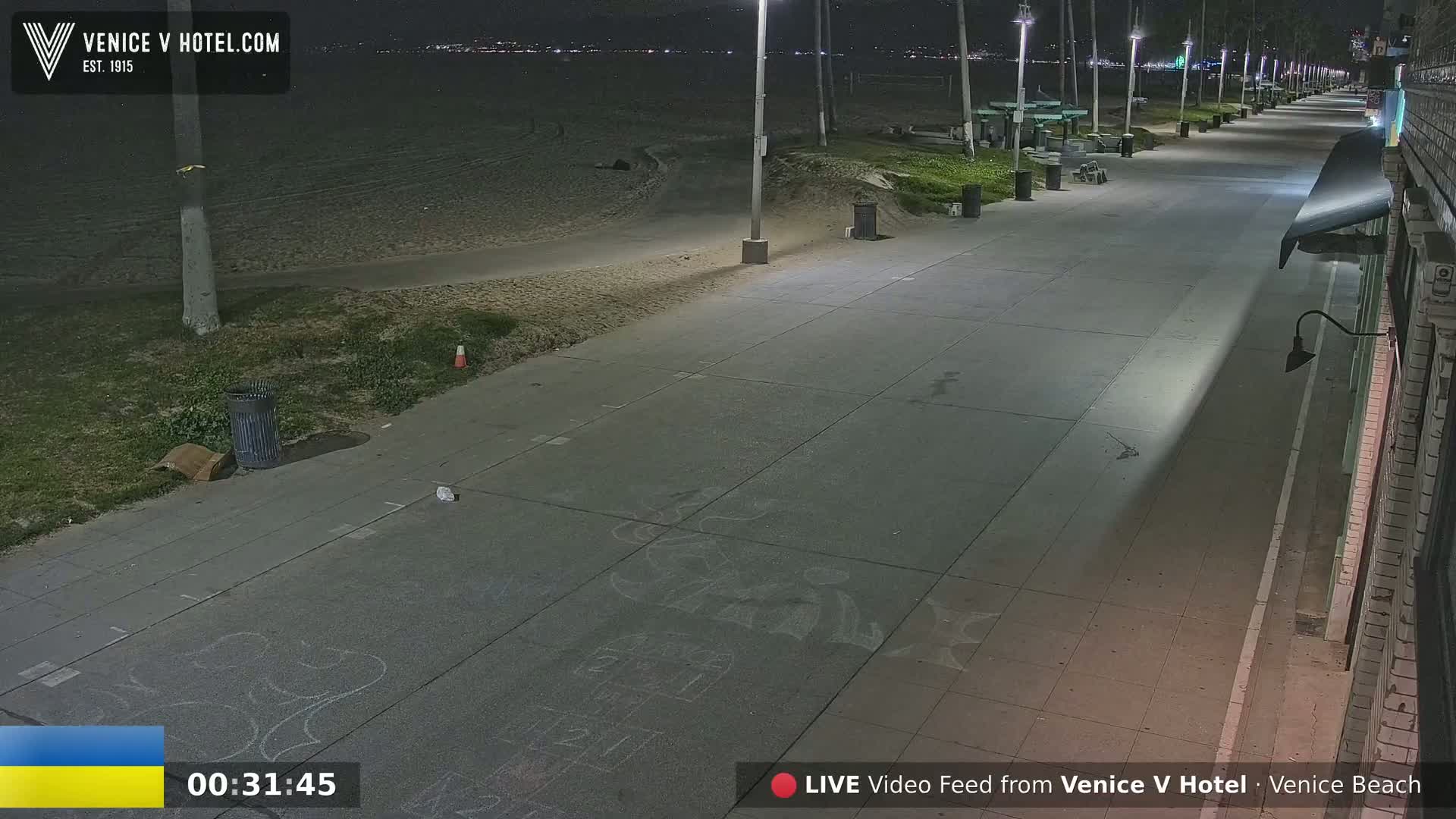 A nighttime view of a mostly empty boardwalk next to a sandy beach, illuminated by streetlights.