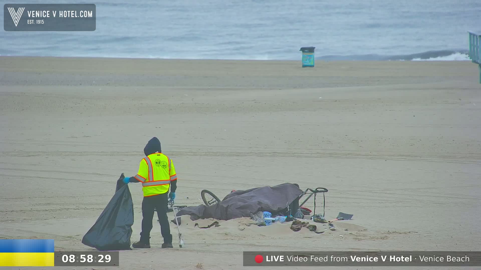 A person in a yellow safety vest collects trash from a sandy beach near a calm ocean.