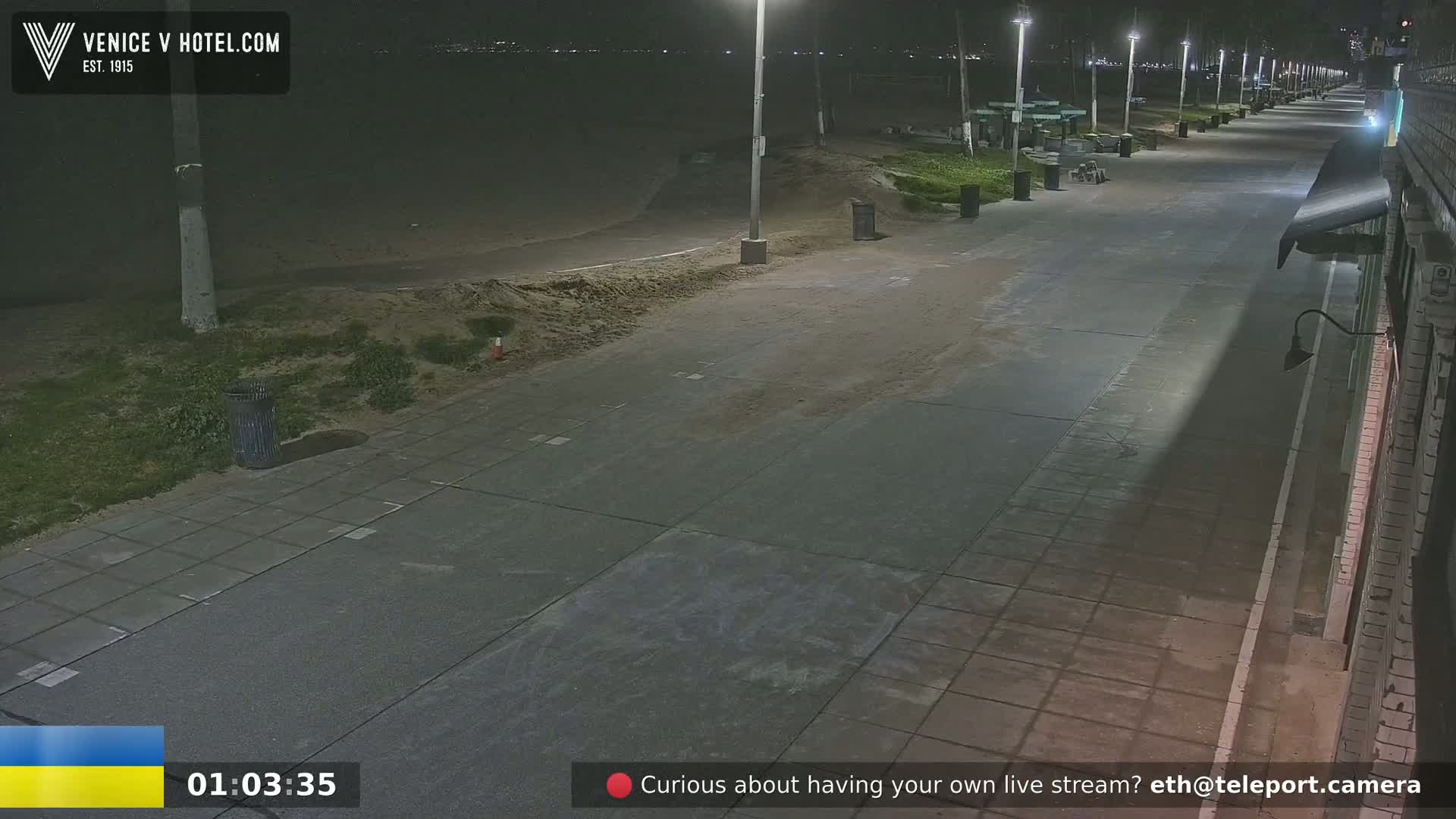 A nighttime view of a mostly empty paved beachfront walkway, with sand and some vegetation to the side, lit by streetlights.