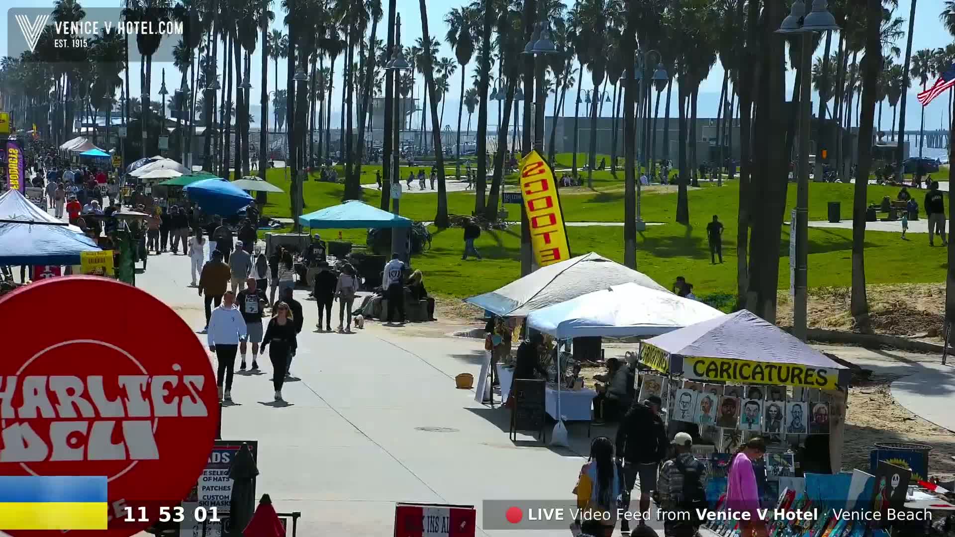 A sunny day at Venice Beach shows many people walking along a boardwalk lined with numerous vendor tents and palm trees.