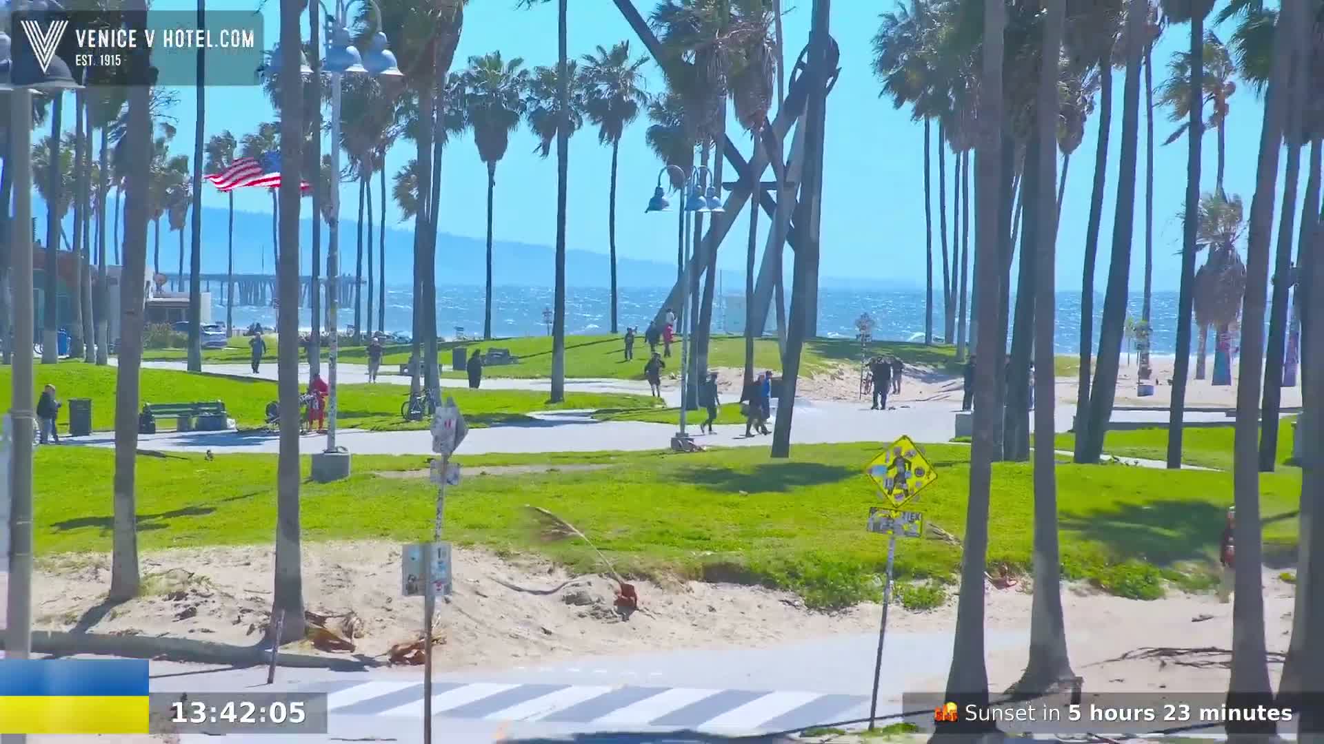 A sunny, windy day at a beachside park with palm trees, a pier in the distance, and people walking along paths.
