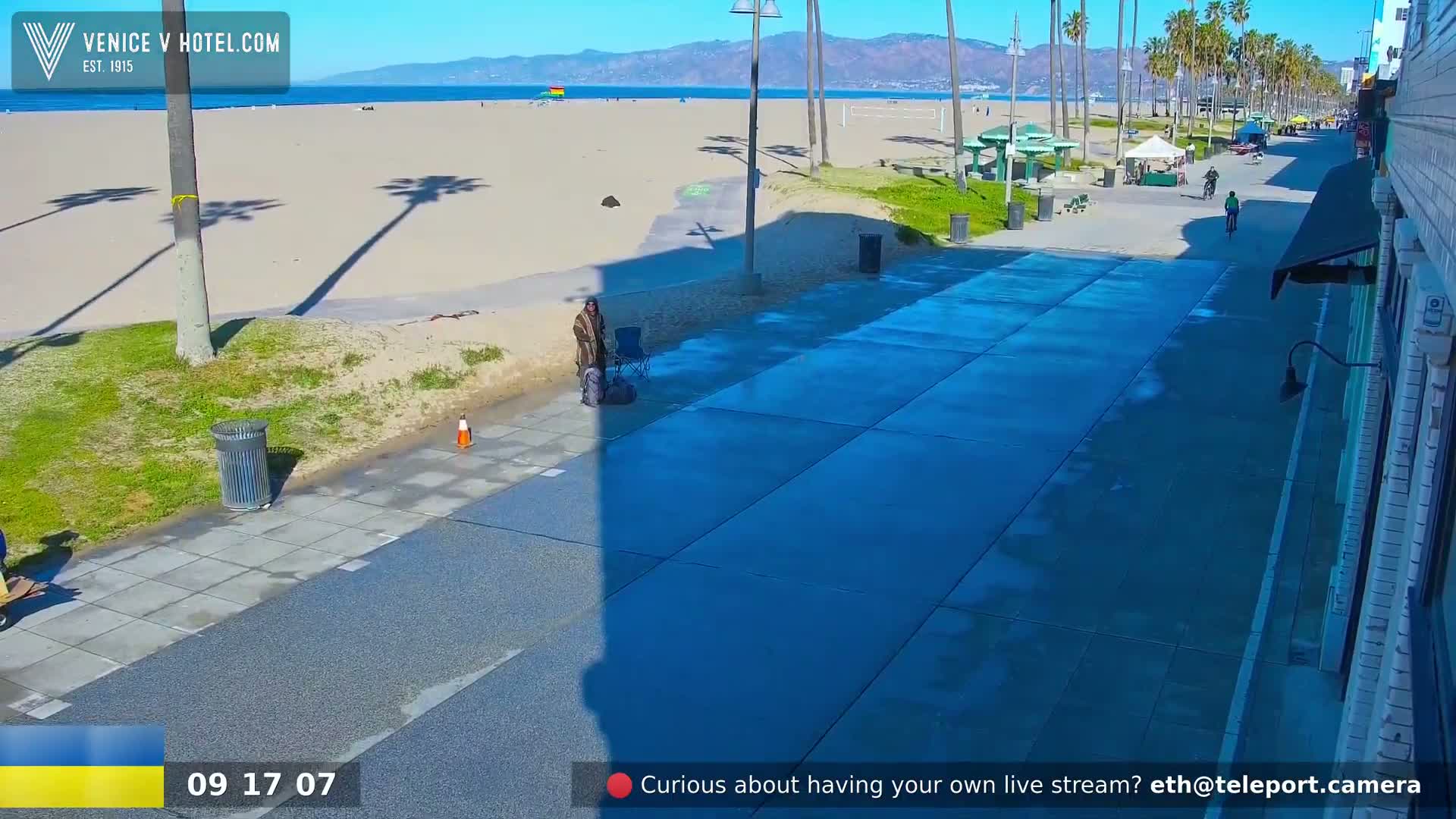 A sunny beach scene is visible from a boardwalk, showing a person sitting on the sidewalk near a trash can and a few people walking or cycling further down the path.