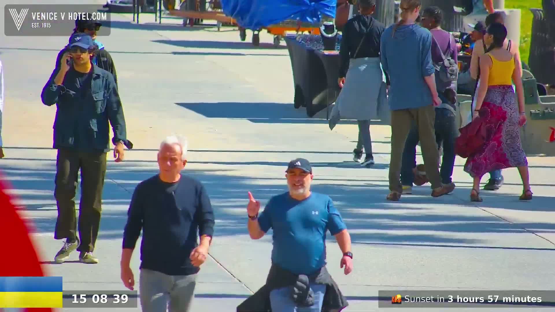Several people walk on a sunny paved area, some appearing to be engaged in conversation, while others walk alone.