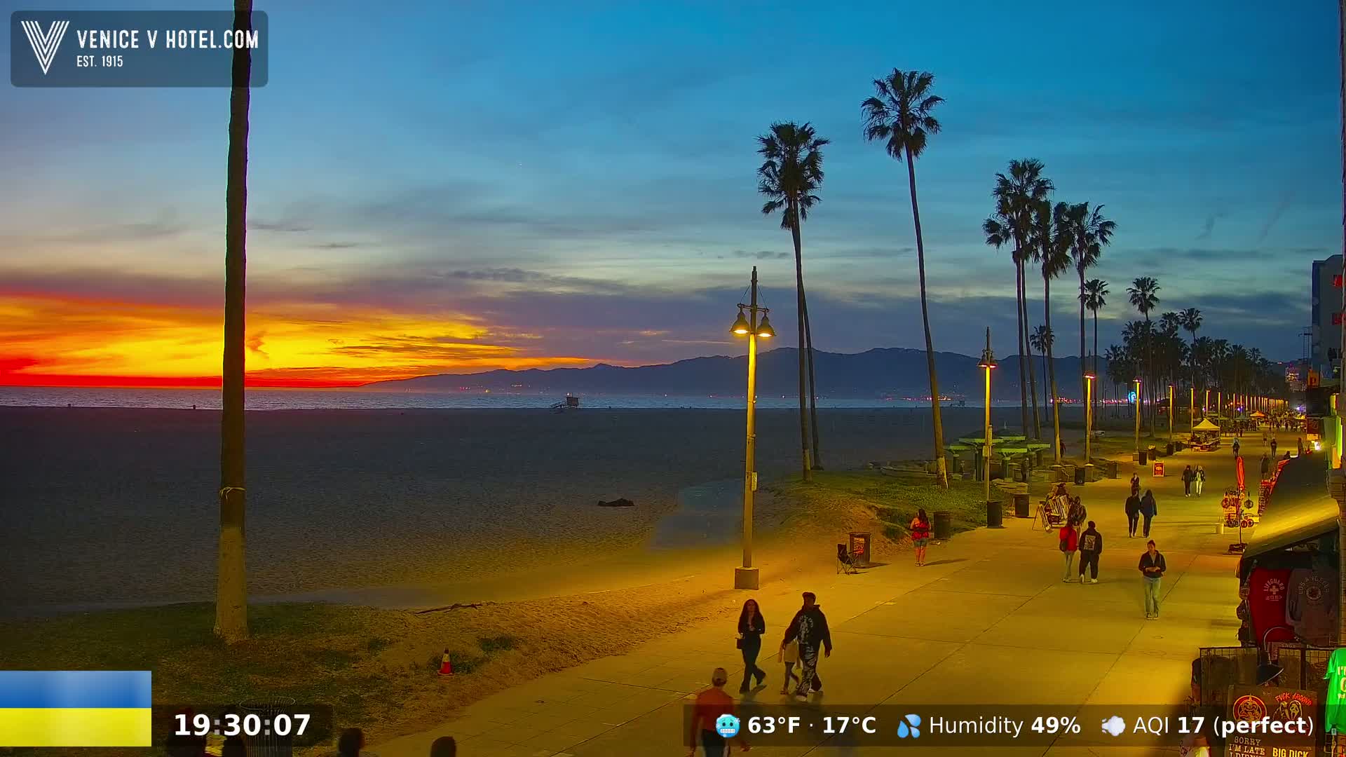 A beach boardwalk at sunset with palm trees, people walking, and a calm ocean under a clear sky.
