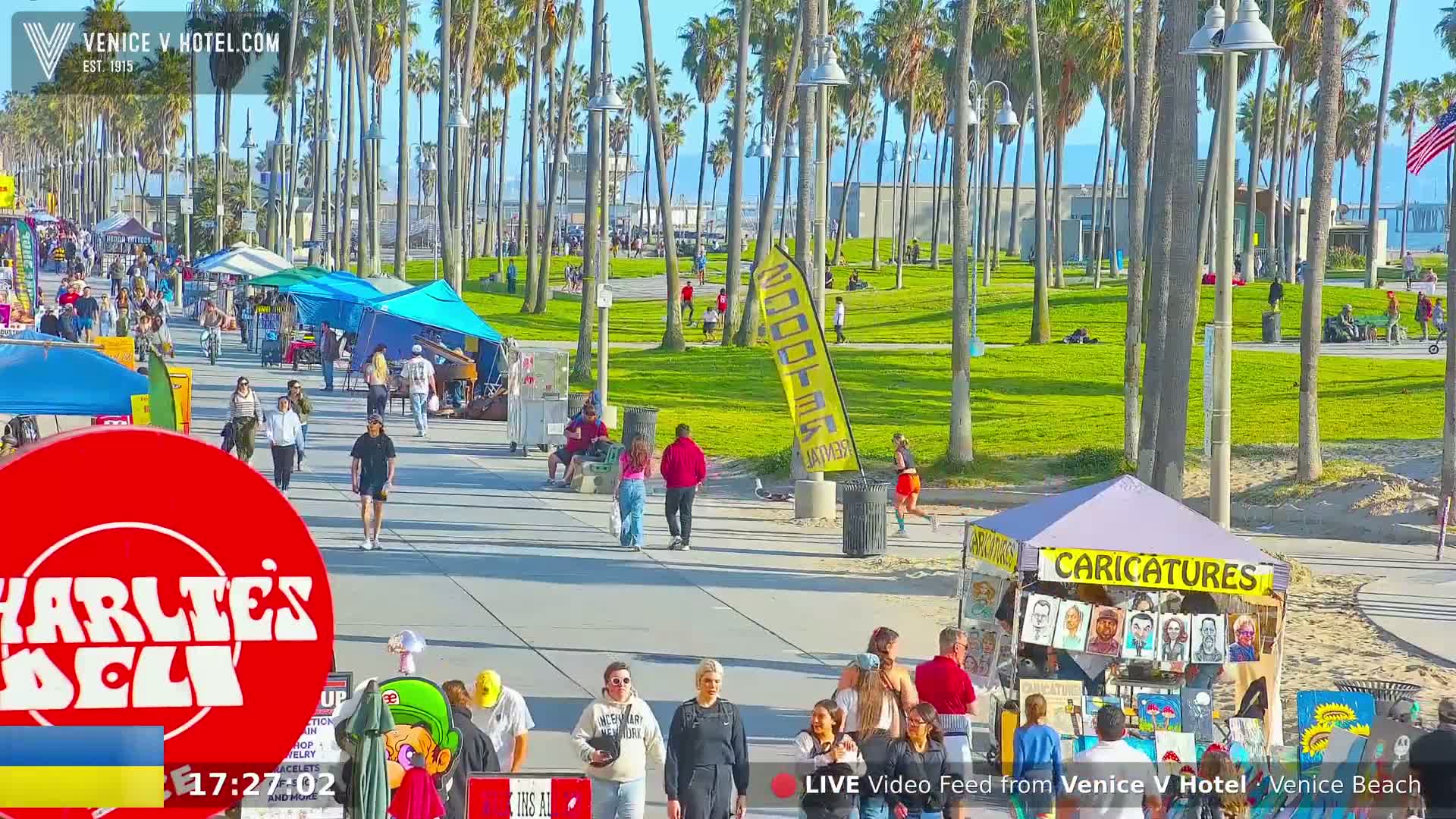 A sunny beach boardwalk scene shows many people walking past various vendor stalls and shops under a bright sky, with palm trees lining the path.