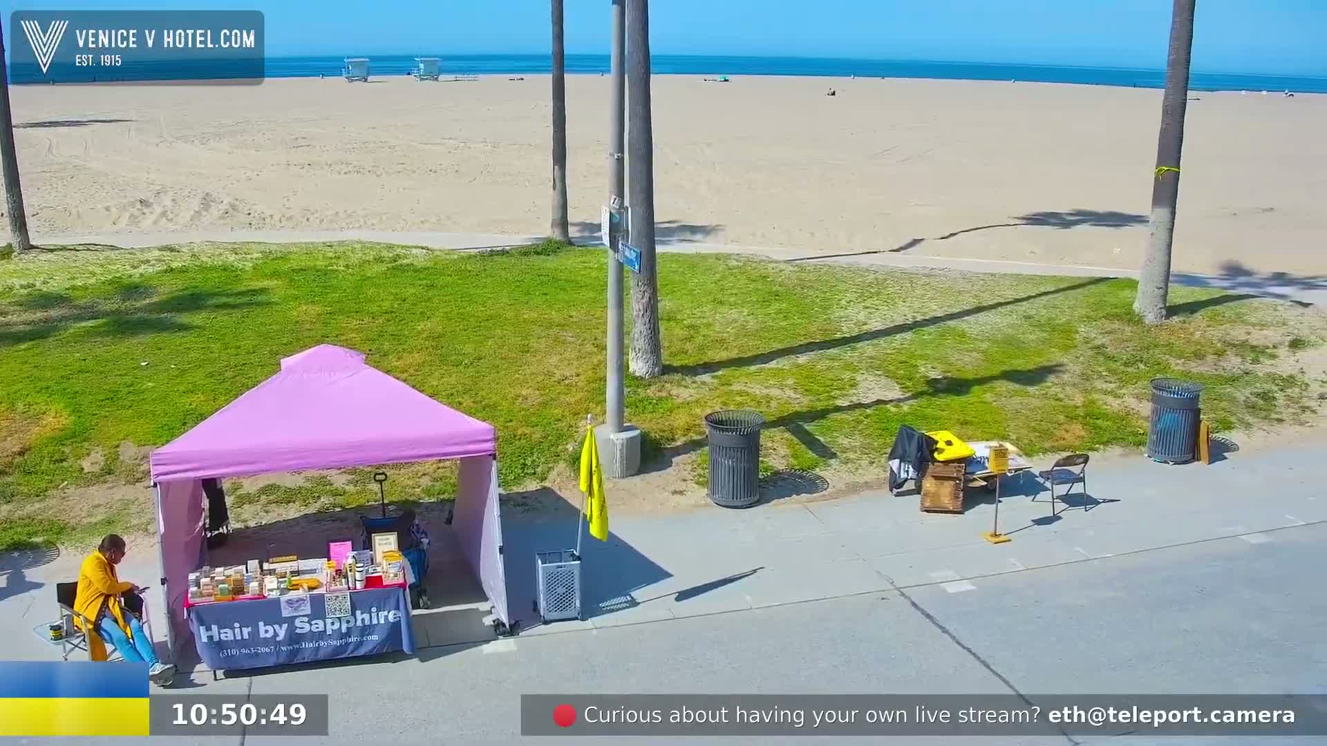 A person sits at a pink vendor tent on a sunny day near a beach with palm trees and trash cans.