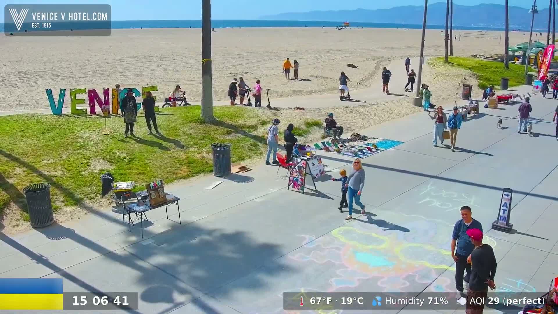 A sunny beach scene shows people walking along a boardwalk near large colorful letters spelling "Venice," with vendors selling artwork and other items, and children drawing on the pavement.