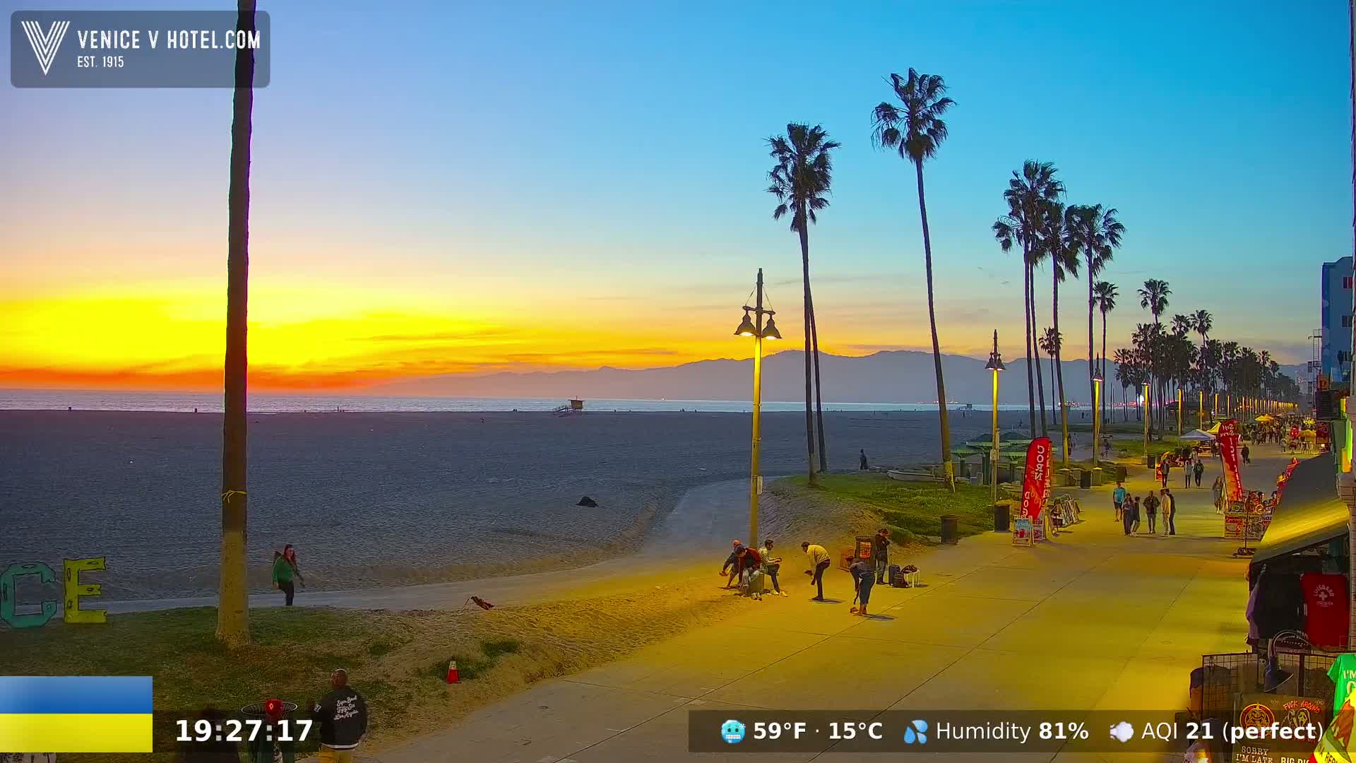 A beach at sunset with palm trees lining a boardwalk where people are walking, under a clear sky.
