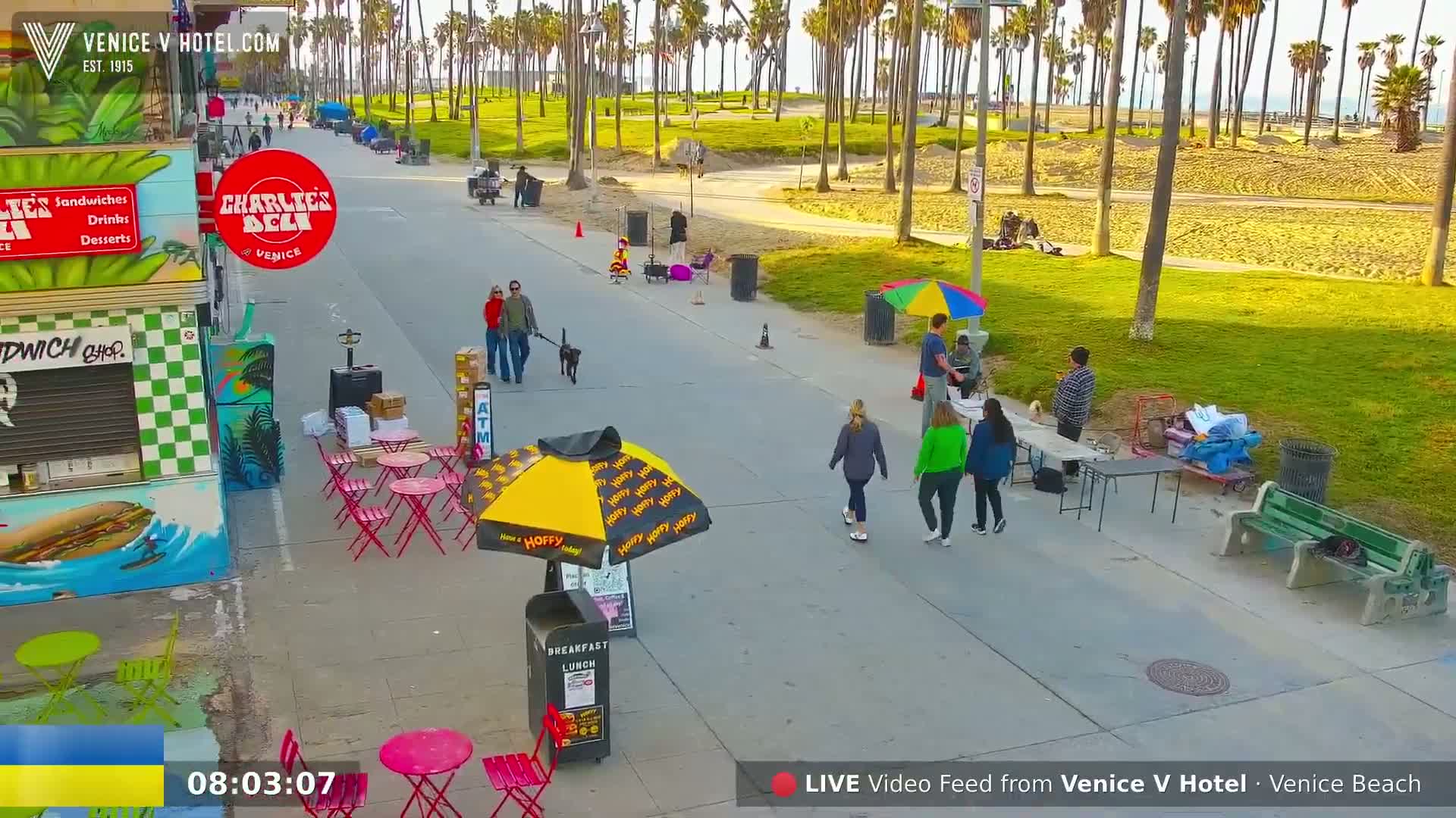 A sunny beachside boardwalk scene shows several people walking, a couple with a dog, and some individuals seated at outdoor tables under umbrellas, near a deli and beachfront grass.