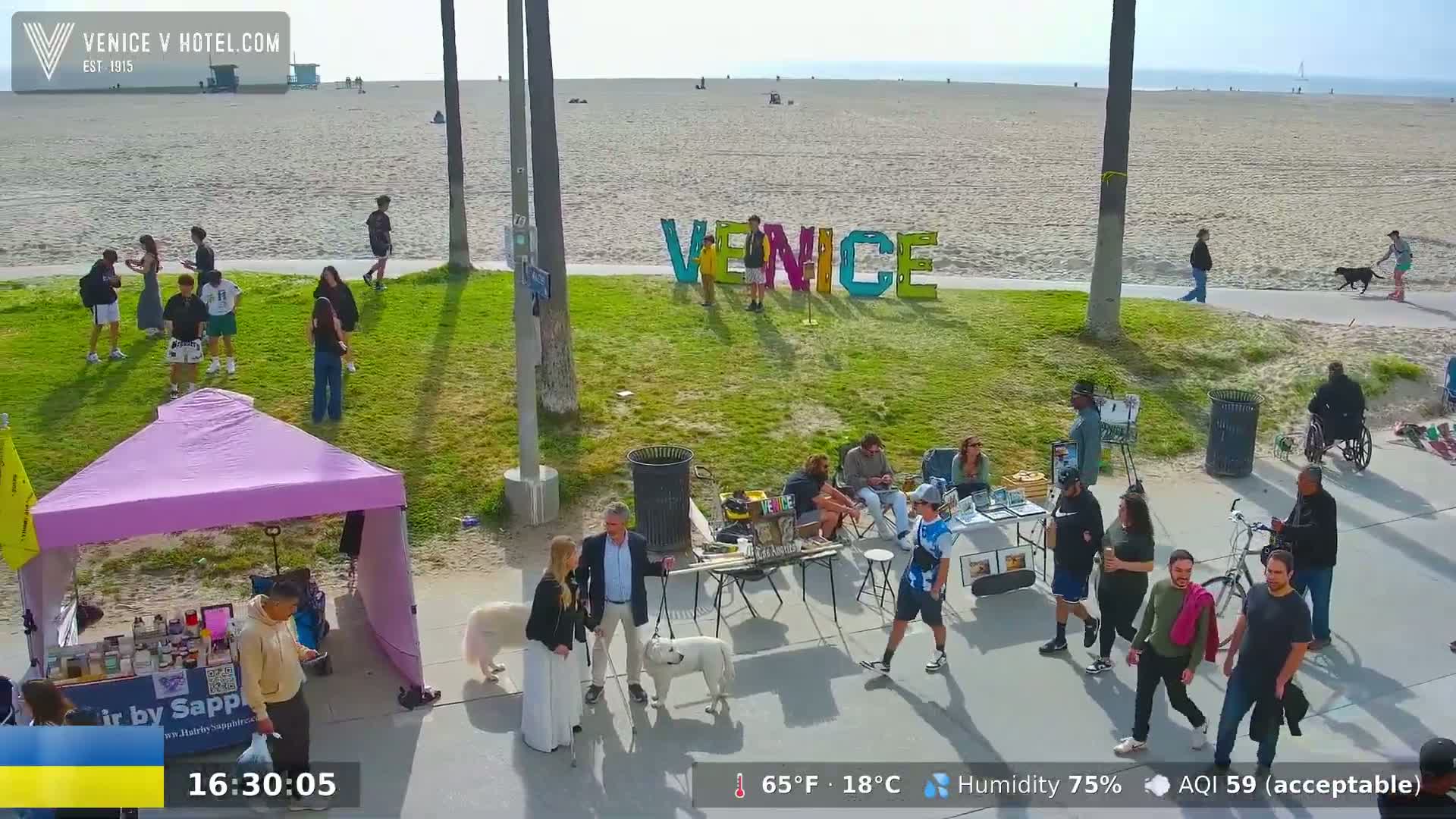 On a sunny day with 75% humidity, people stroll along a beachside path near a large "VENICE" sign, with several small vendors and their wares set up on the grass.