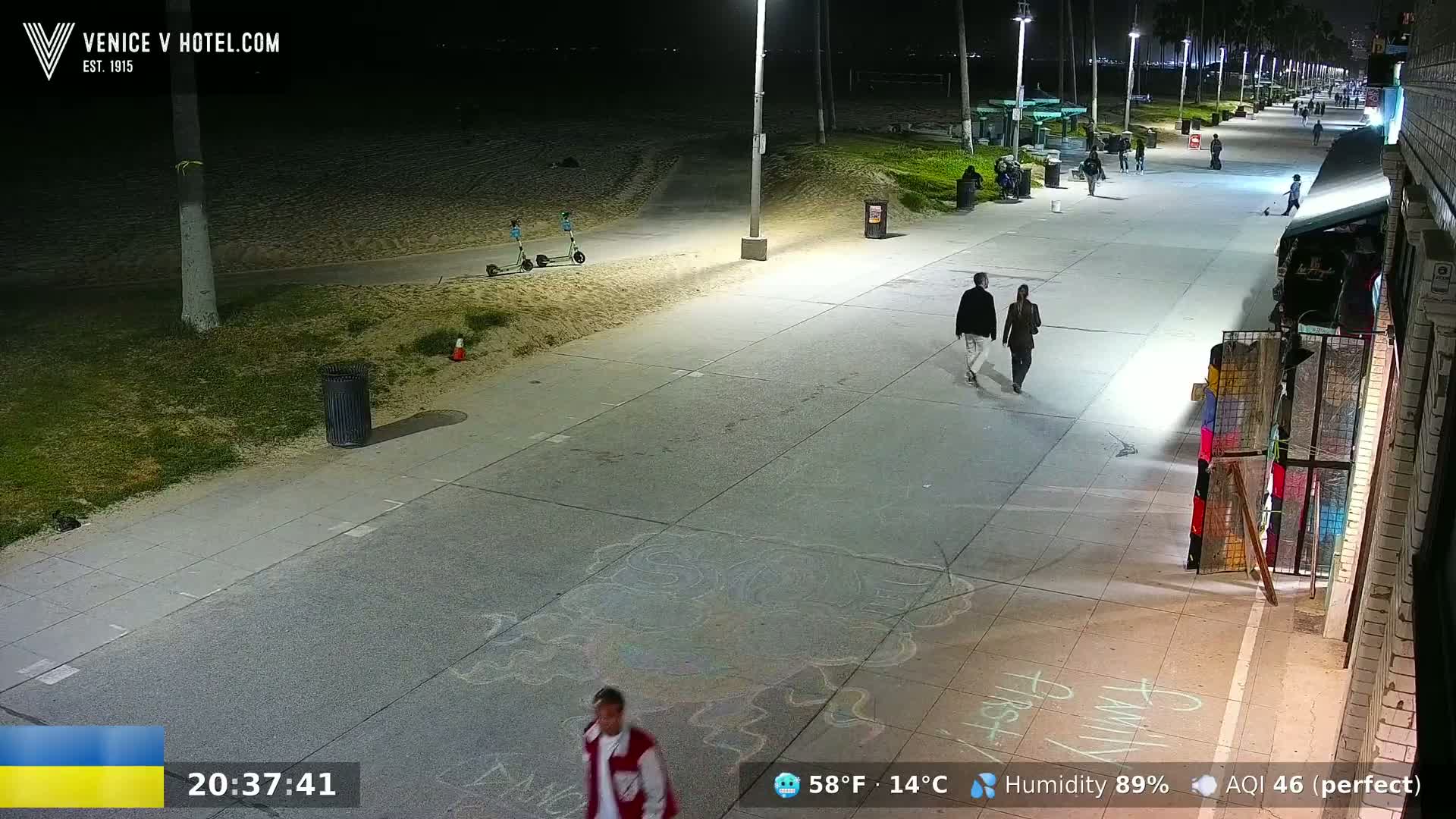 A nighttime view of a paved beachfront walkway with several people walking, two electric scooters parked on the sand, and chalk drawings on the ground.