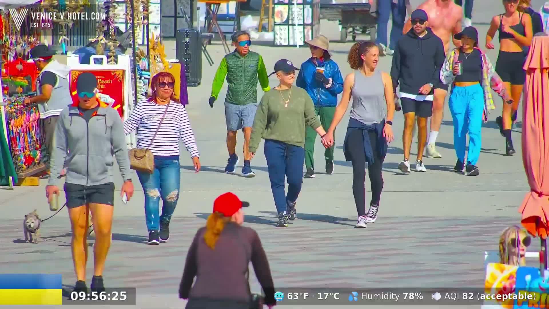 A sunny day shows many people walking on a paved walkway near various shops and stands, one person walking a small dog.
