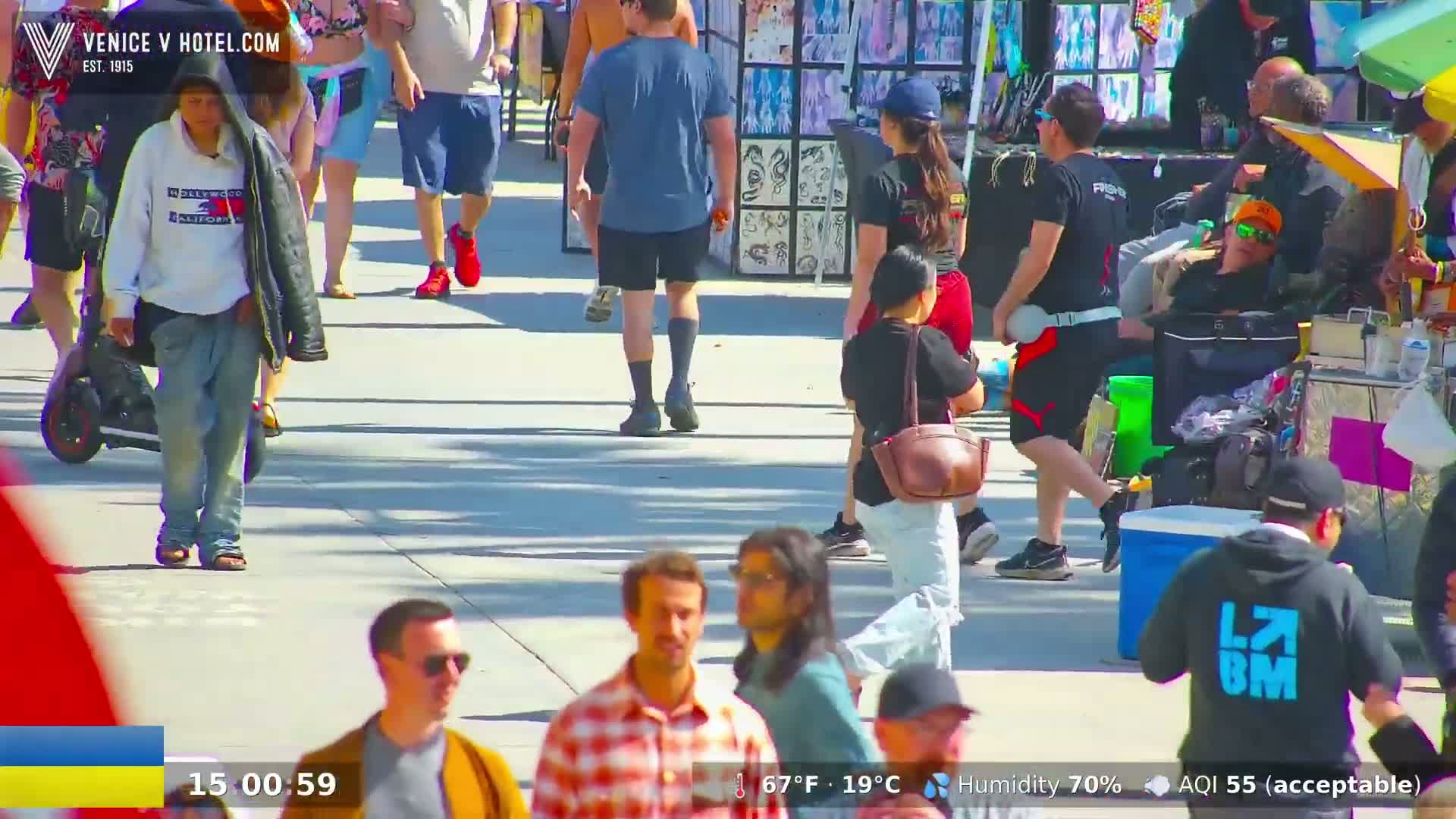 A sunny day with a humidity of 70% shows many people walking on a paved walkway, some near various stalls and vendors.