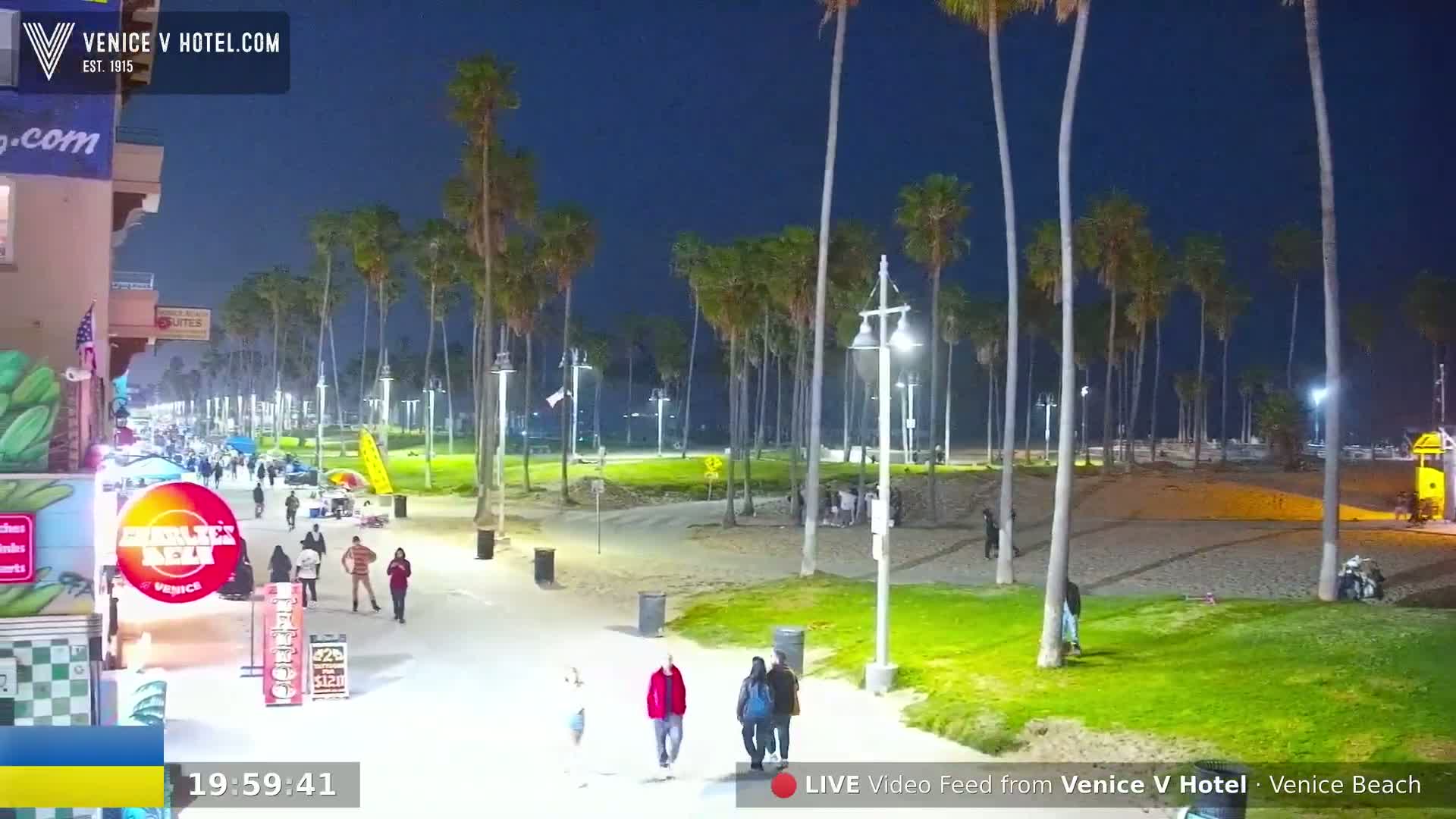 A nighttime view of a beach boardwalk with palm trees, people walking, and small businesses along the side, under a clear, dark blue sky.