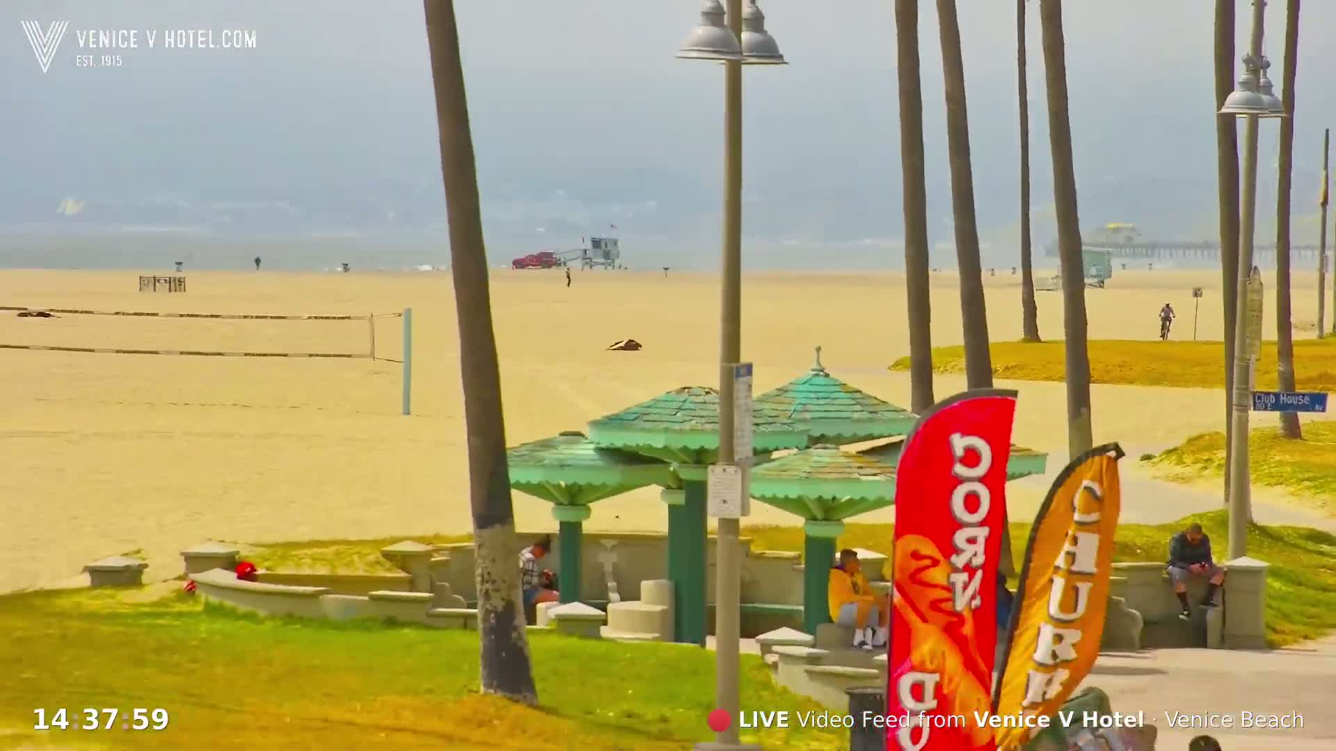 A hazy, sunny day at a beach shows sandy shores, palm trees, beach umbrellas, and several people relaxing near a volleyball net and lifeguard tower in the distance.