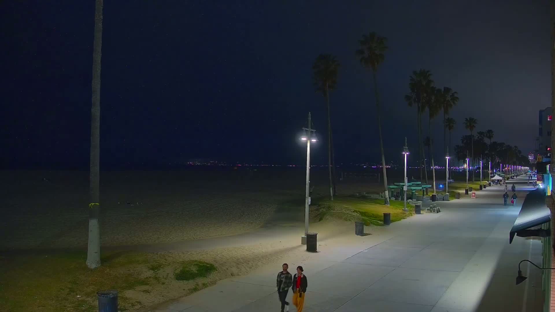 A nighttime beach boardwalk scene shows two people walking, palm trees lining the path, and a sandy beach under a clear, starry sky.