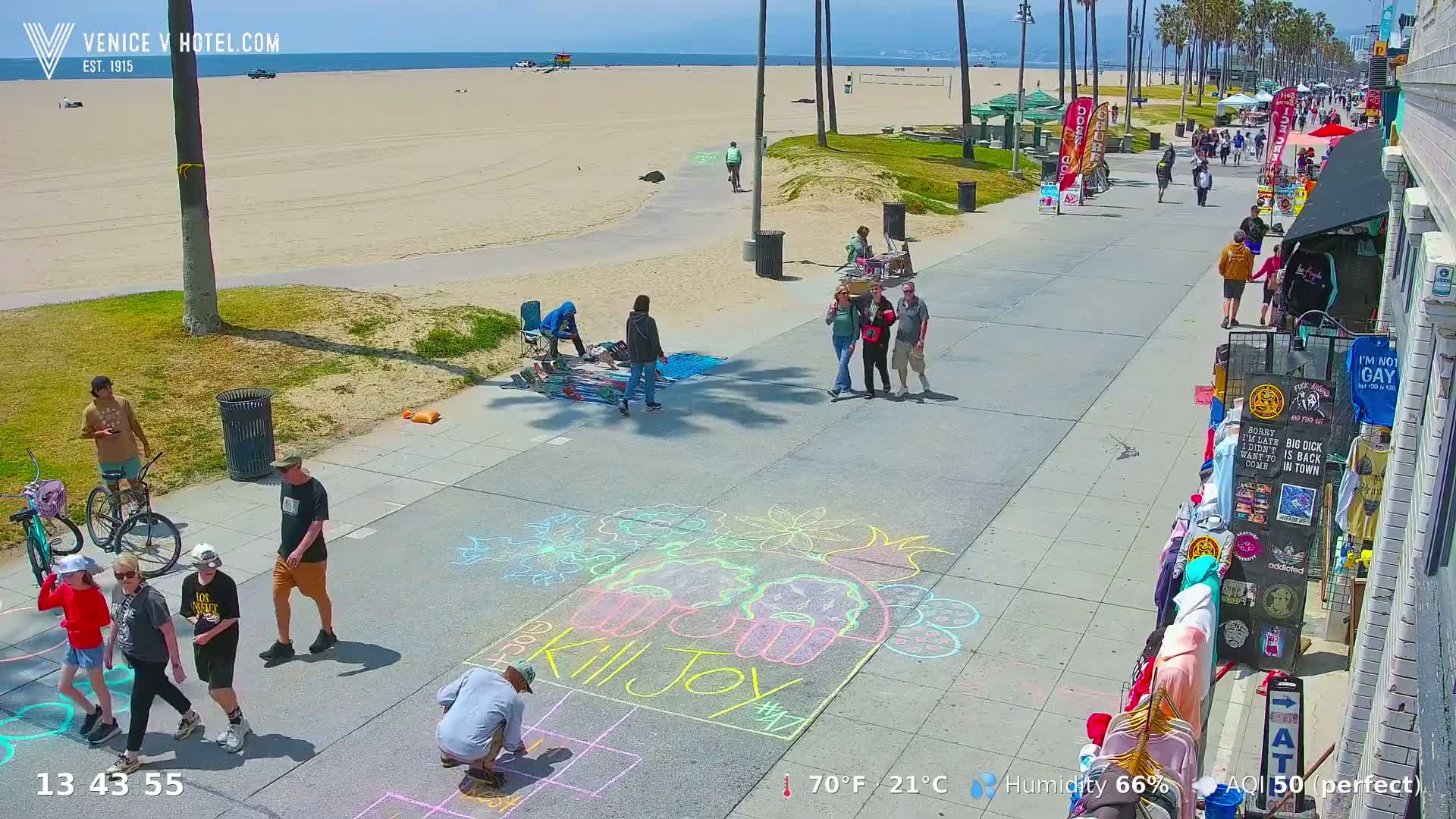 A sunny beach boardwalk scene shows people walking, vendors selling goods, and chalk art on the pavement.
