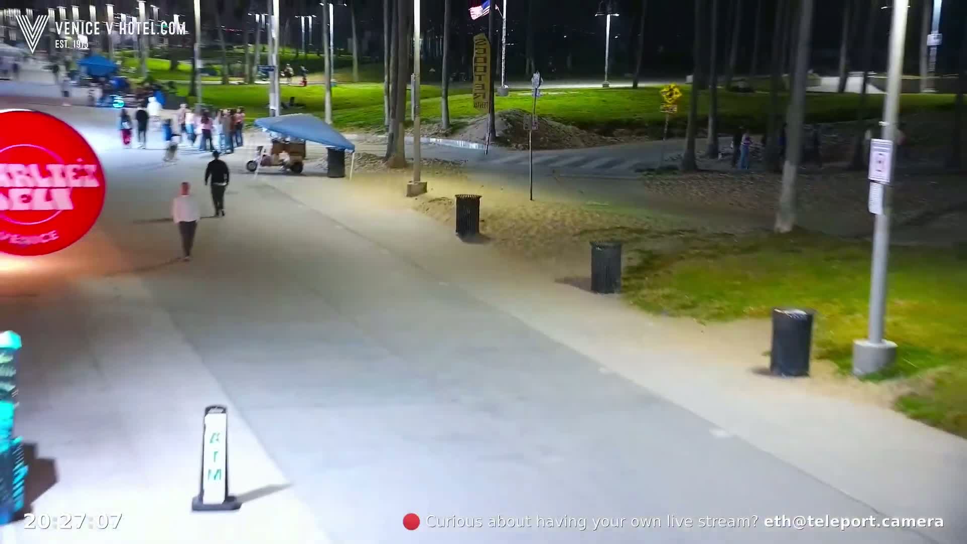 A nighttime scene shows a paved walkway with several pedestrians, trash cans, and a large illuminated circular sign; a grassy area and trees are visible in the background.