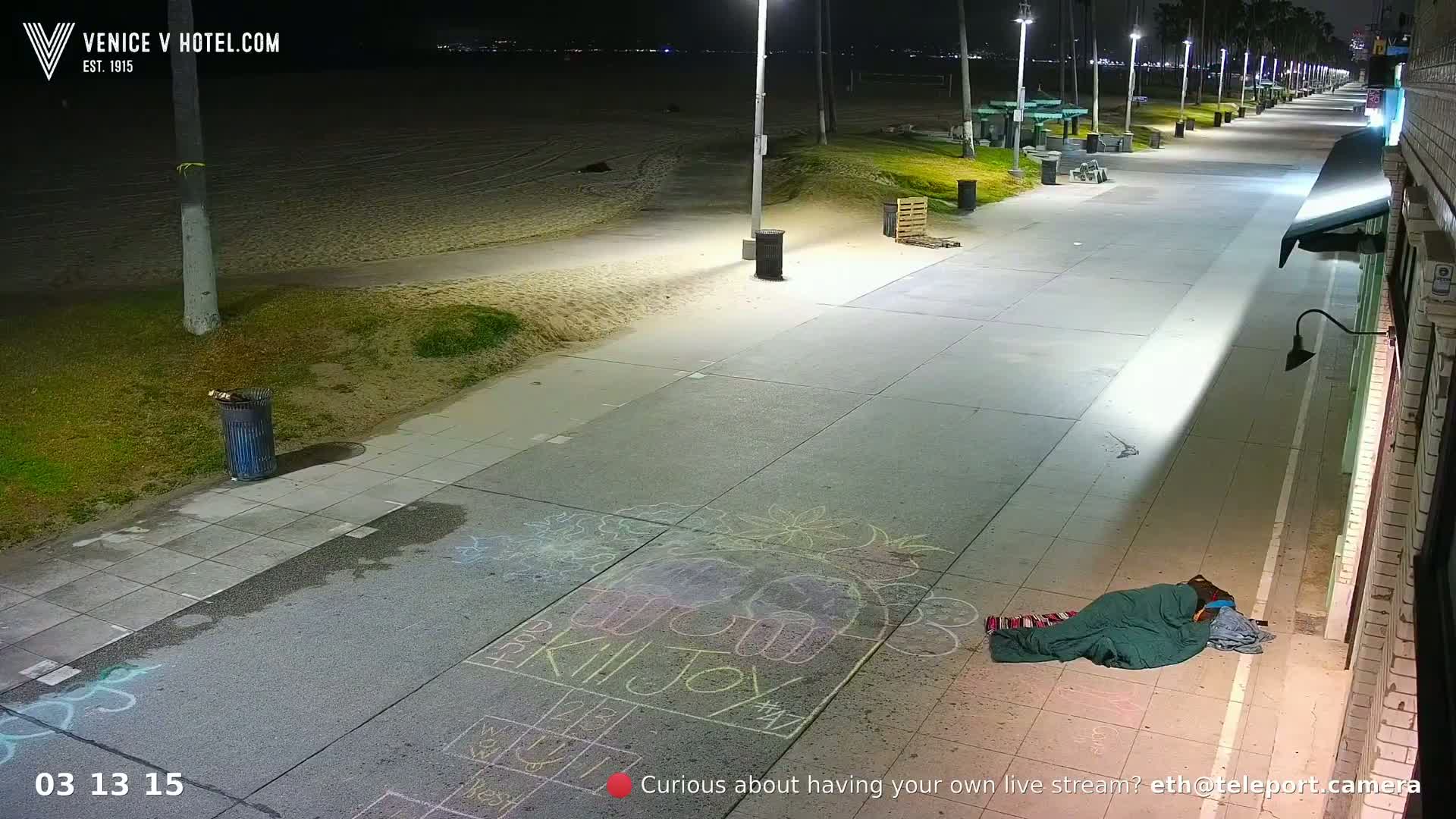 A person sleeps under a blanket on a paved walkway at night, next to children's chalk drawings, near a beach under bright lights.