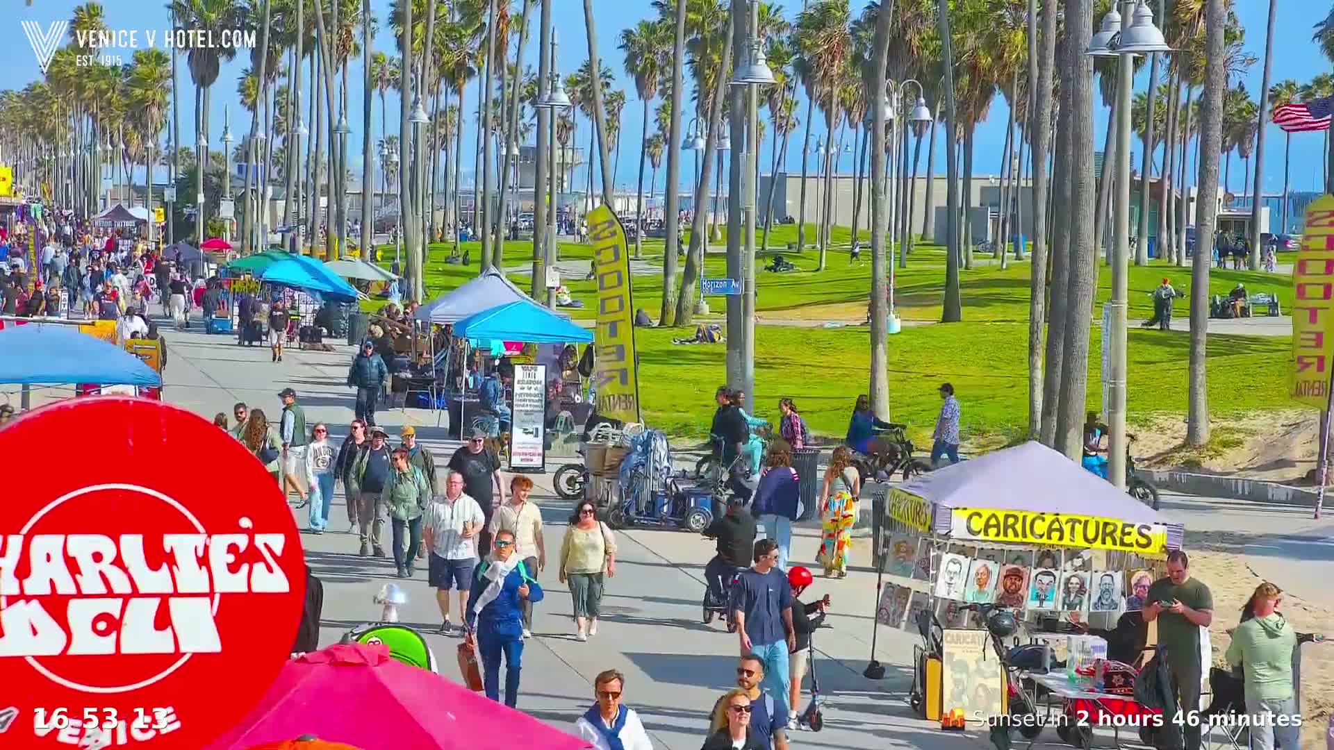 A sunny boardwalk scene shows many people walking among numerous small vendors and stands under a bright sky, with palm trees lining a grassy area in the background.