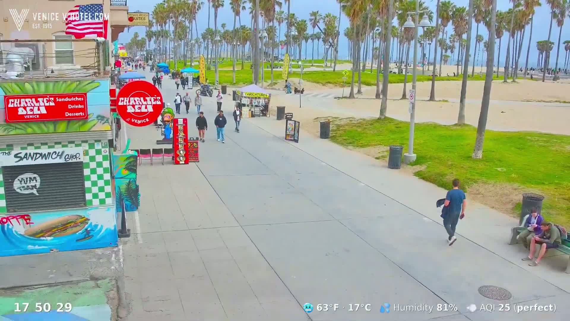 A sunny boardwalk scene shows pedestrians walking past shops and toward a sandy beach lined with palm trees.