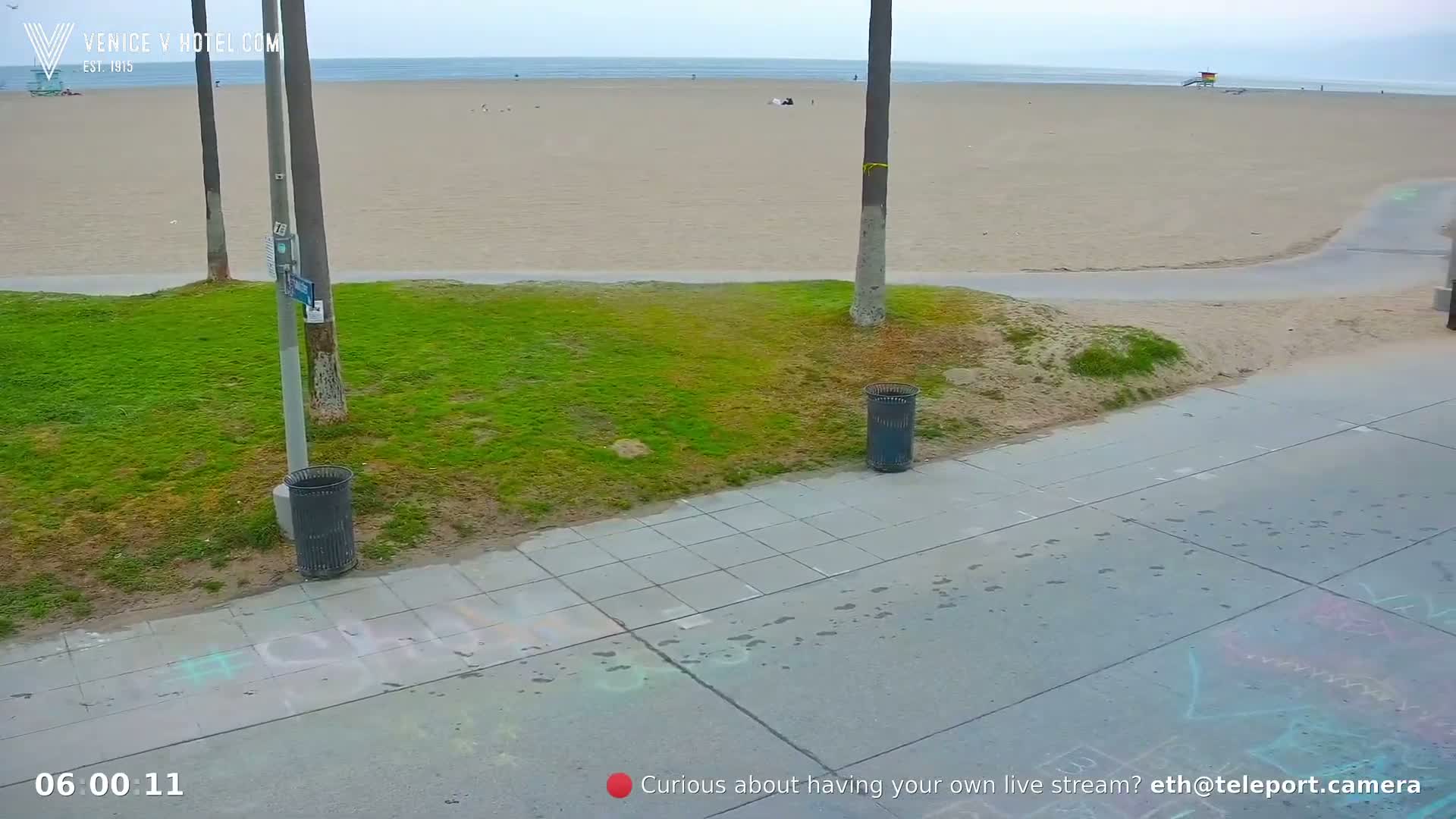 A mostly empty sandy beach is visible beyond a grassy area with trash cans and a paved walkway showing footprints and chalk drawings.
