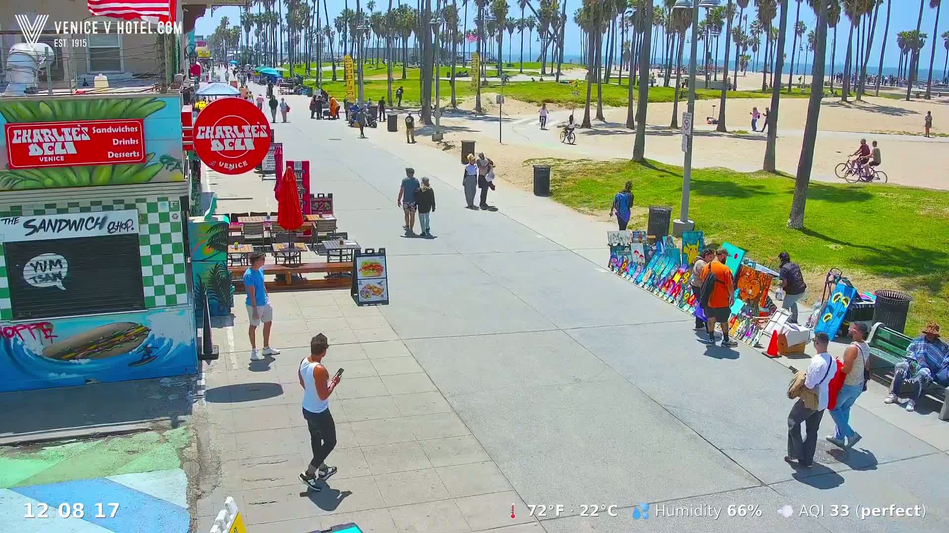 A sunny boardwalk scene shows pedestrians strolling past vendors selling artwork and a deli, with a beach and ocean in the background.