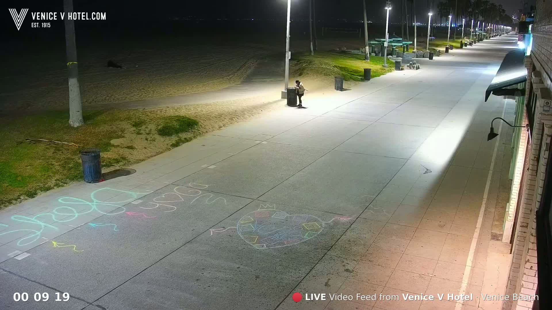 A nighttime view of a paved walkway next to a sandy beach, illuminated by streetlights, with chalk drawings on the ground and a lone person standing near a trash can.
