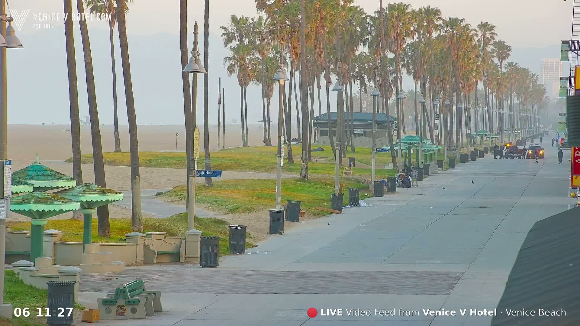 A hazy morning view shows a beach boardwalk lined with palm trees, benches, trash cans, and a few people walking, with a sandy beach and ocean visible in the distance.