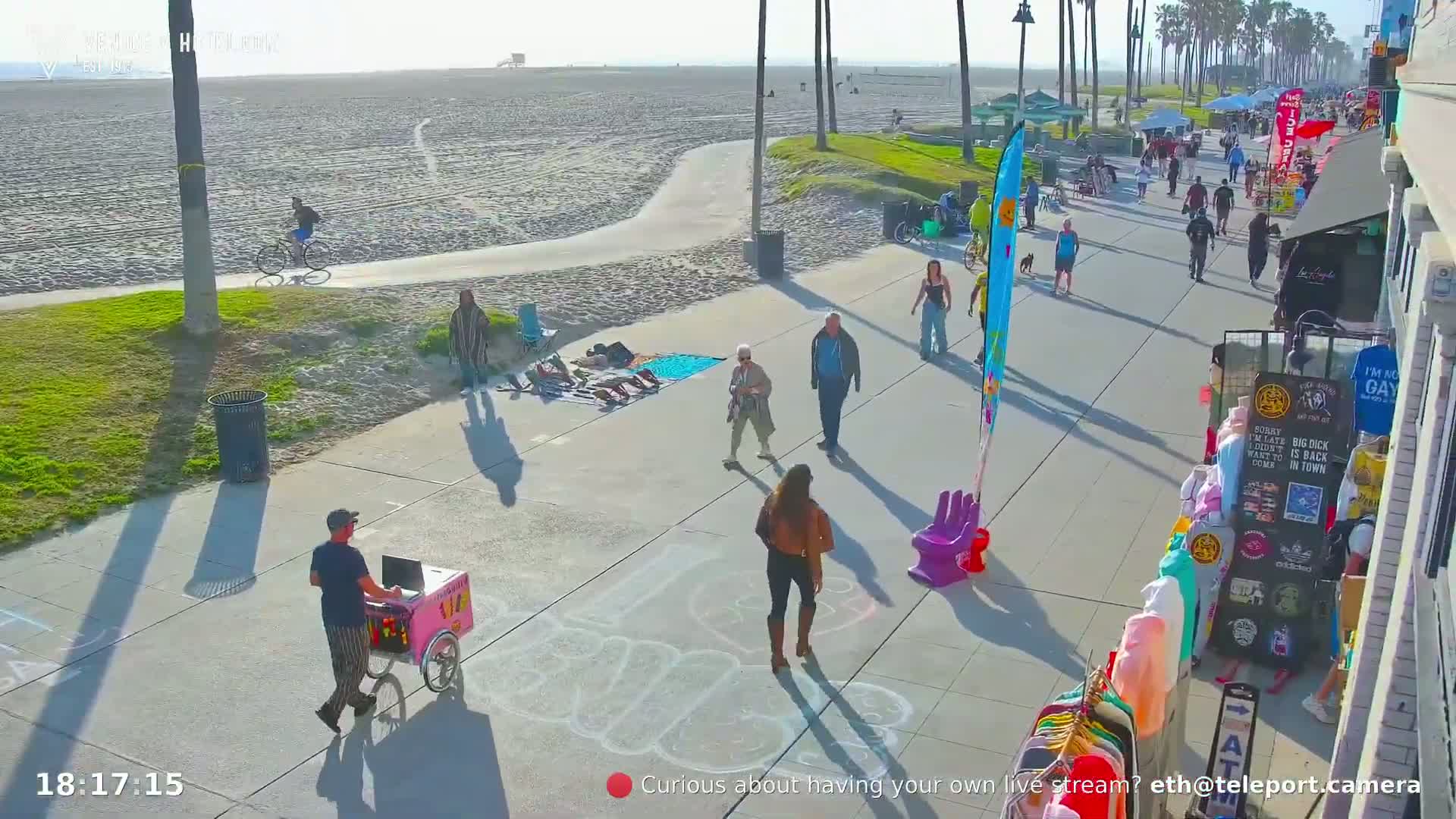 A sunny beach boardwalk scene shows people walking, biking, and selling goods, with a sandy beach and ocean in the background.