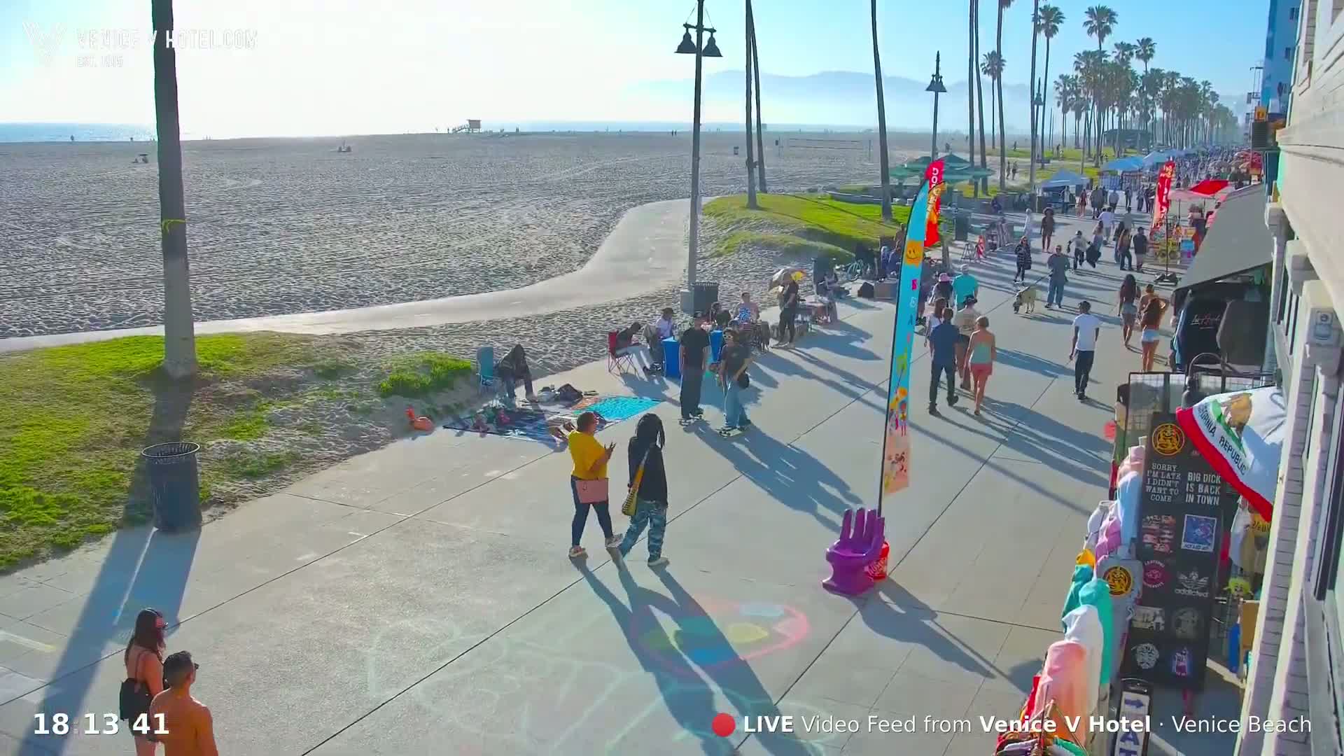 A sunny beach boardwalk scene shows many people walking, some vendors selling goods, and a sandy beach with the ocean in the background.
