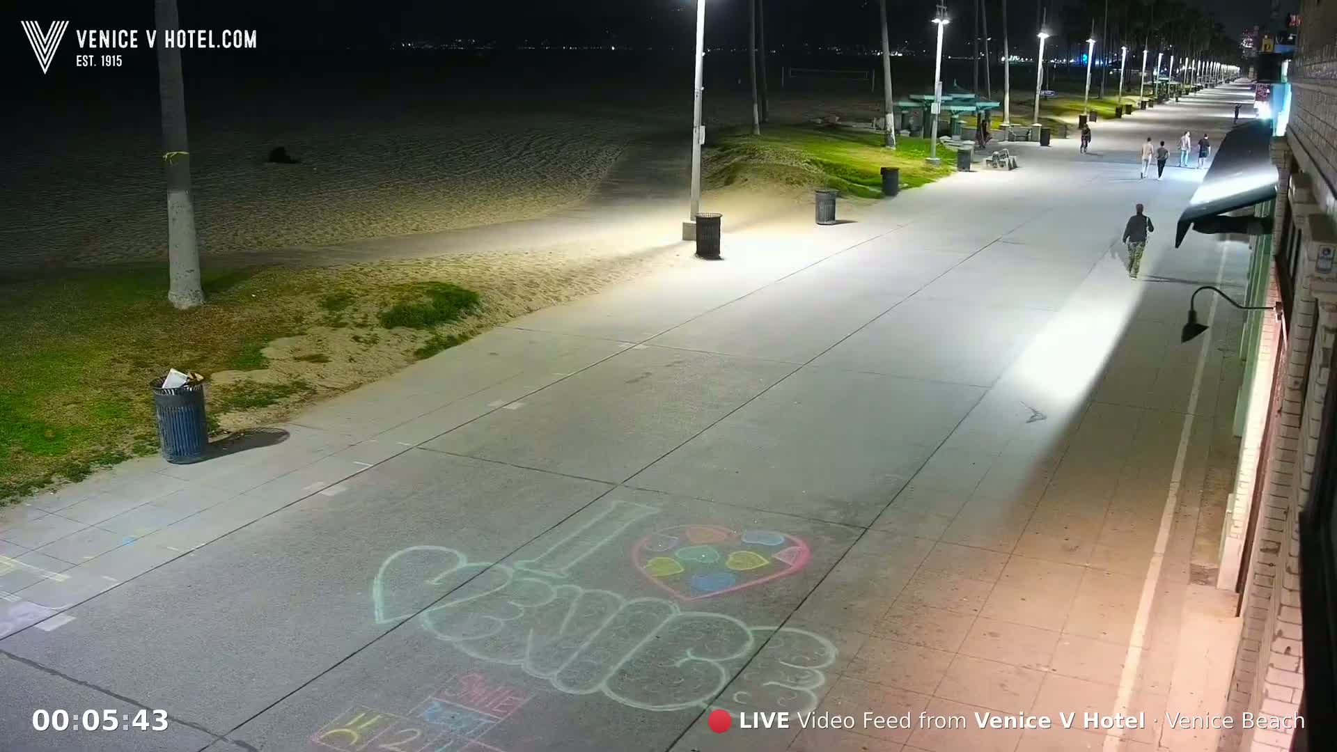 A nighttime view of a paved beach boardwalk with several people walking, illuminated by streetlights, next to a sandy beach.