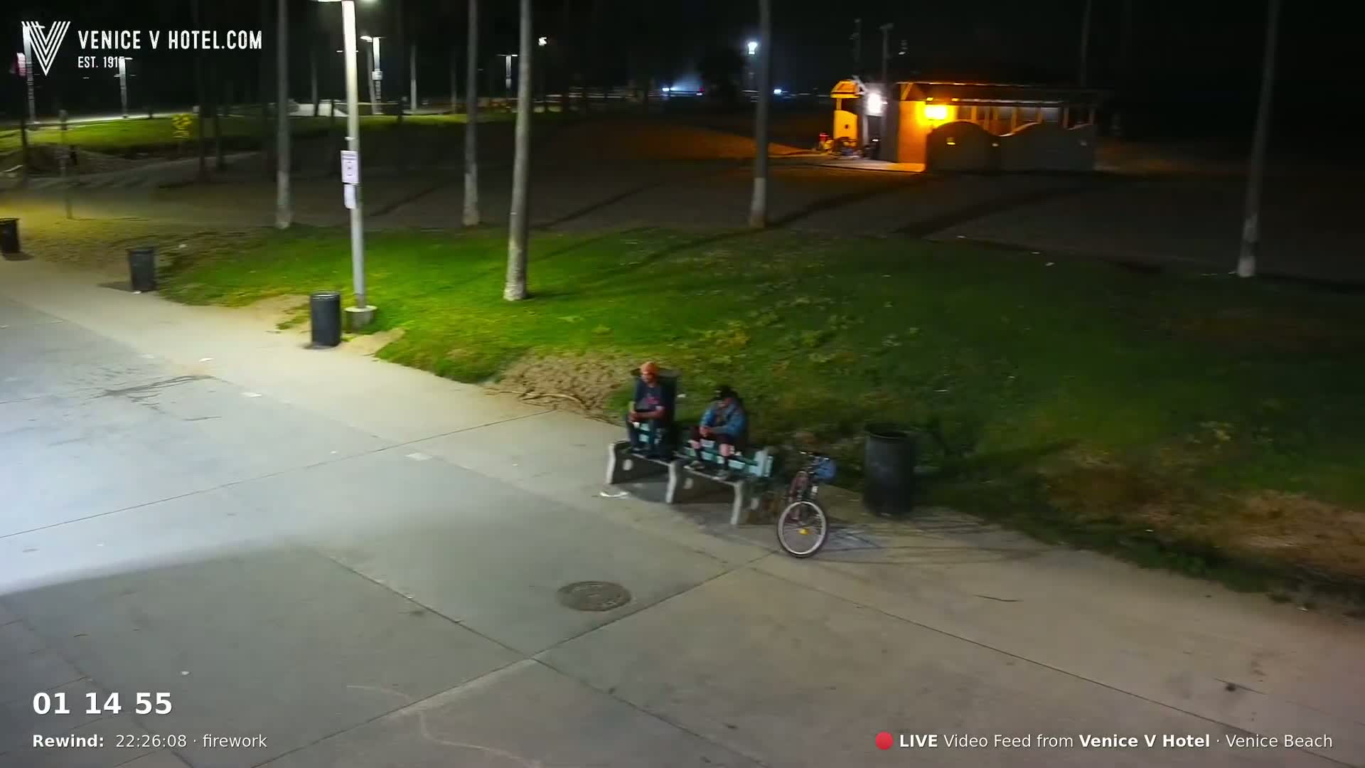 Two people sit on benches at night next to a bicycle, near a grassy area and a building, under artificial light.