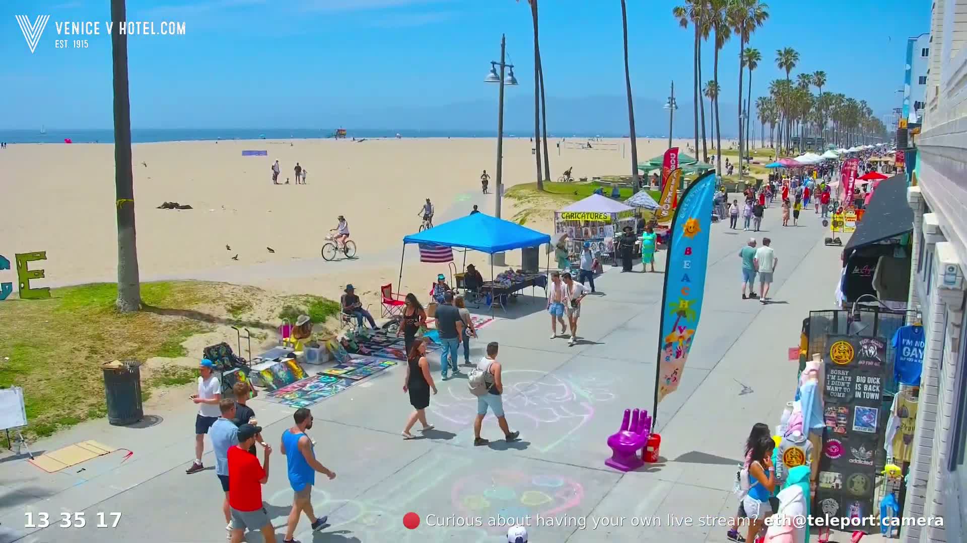 A sunny beach boardwalk scene shows numerous people strolling, vendors selling goods under tents, and artists displaying artwork on the paved walkway, next to a sandy beach with a few people on it.