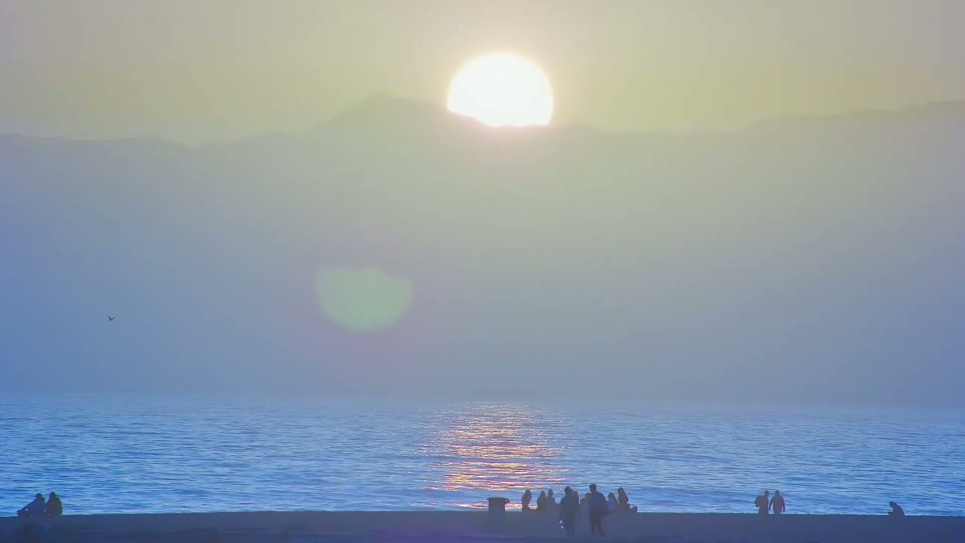 A hazy sunset over a calm ocean with a few people silhouetted on a beach in the foreground.