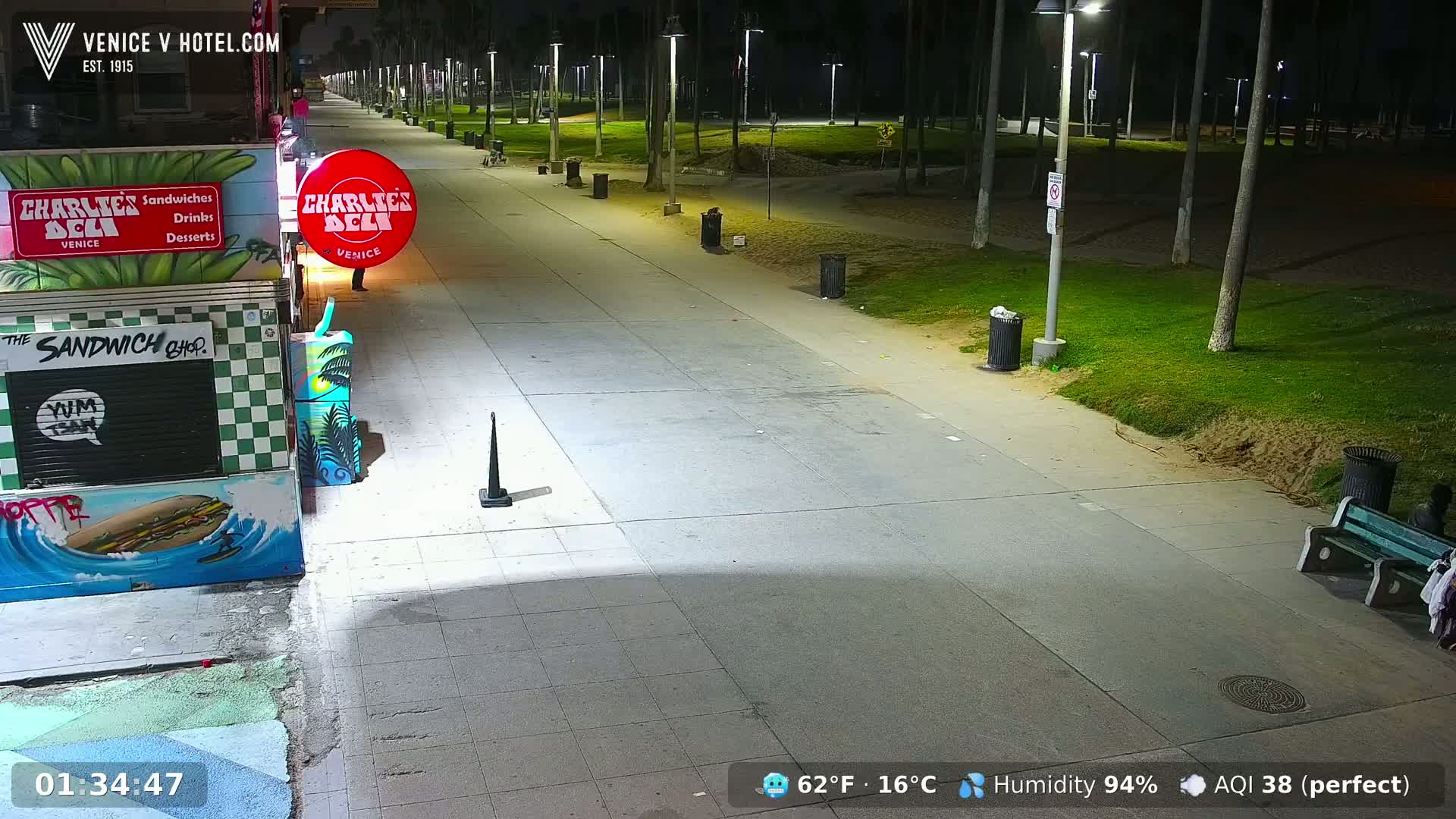 A nighttime view of a mostly empty boardwalk with palm trees, grassy areas, and a few trash cans and benches under bright streetlights, at 62°F and 94% humidity.