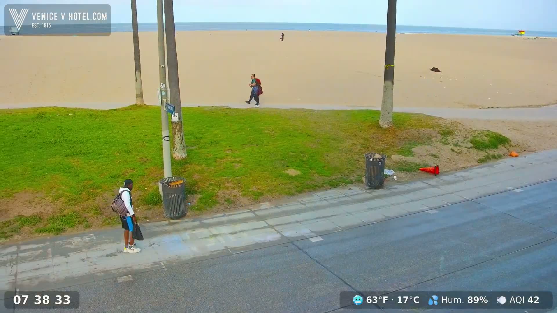 A sandy beach with a calm ocean stretches behind a grassy area, where two people walk along a sidewalk beside trash cans and palm trees under a partly cloudy sky.