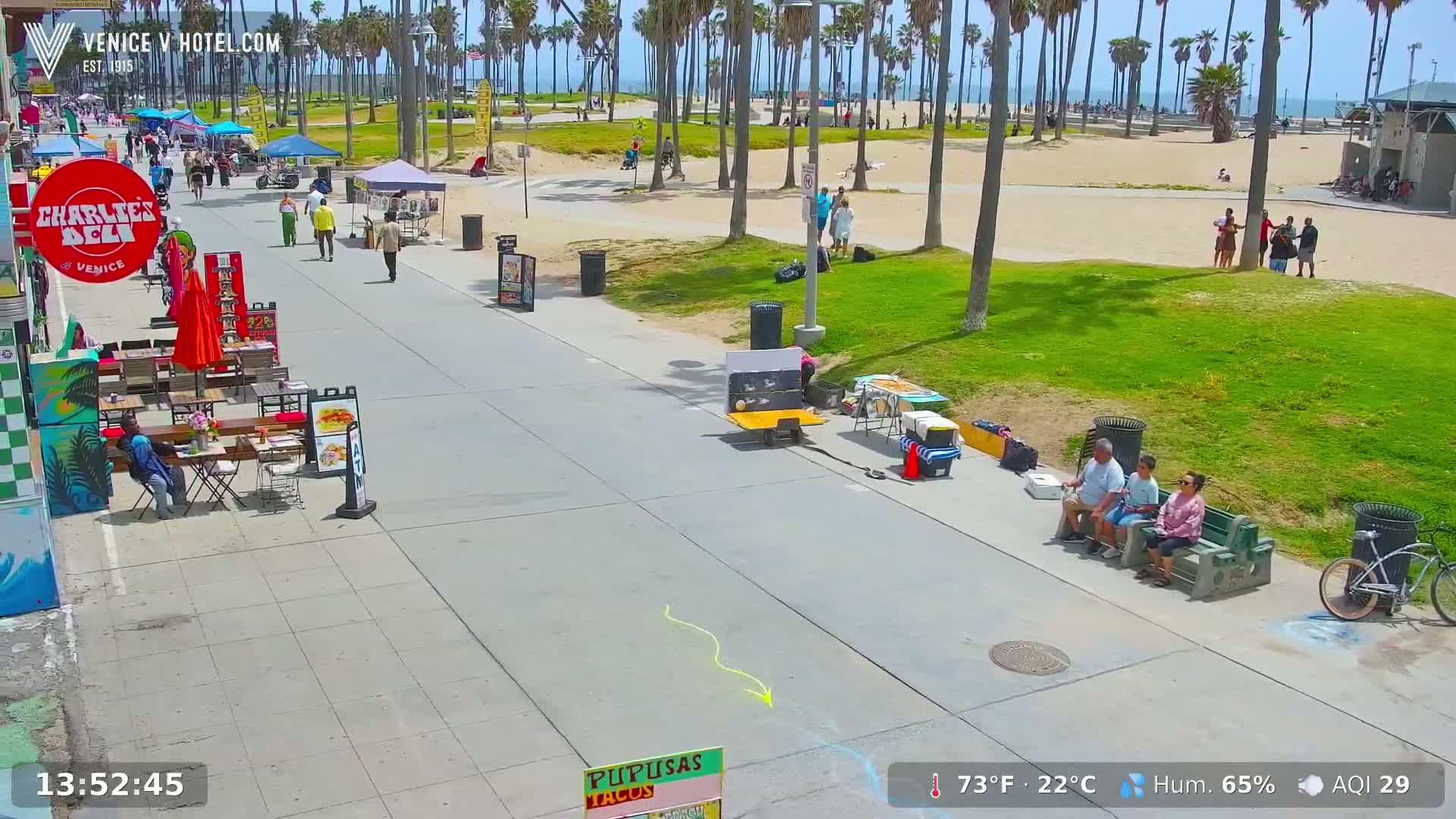 A sunny boardwalk scene shows people walking and sitting near food vendors and a beach with palm trees under a clear sky.
