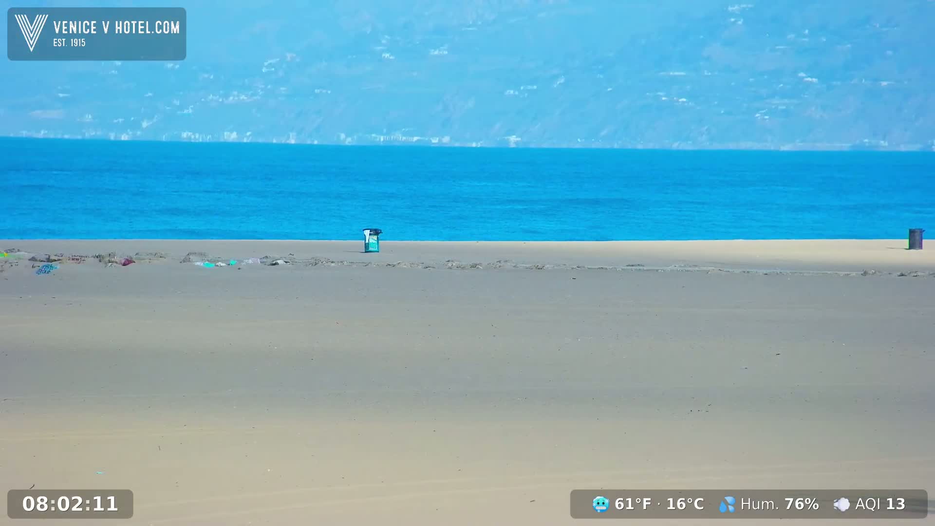 A mostly empty sandy beach with a trash can and some scattered debris stretches to a bright blue ocean under a clear sky.