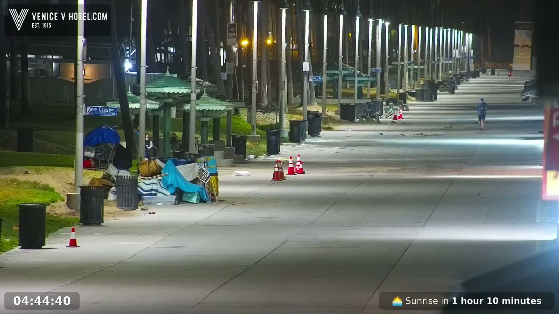 A nighttime scene shows a mostly empty boardwalk with several trash cans and some homeless belongings near park benches, and a lone jogger in the distance.
