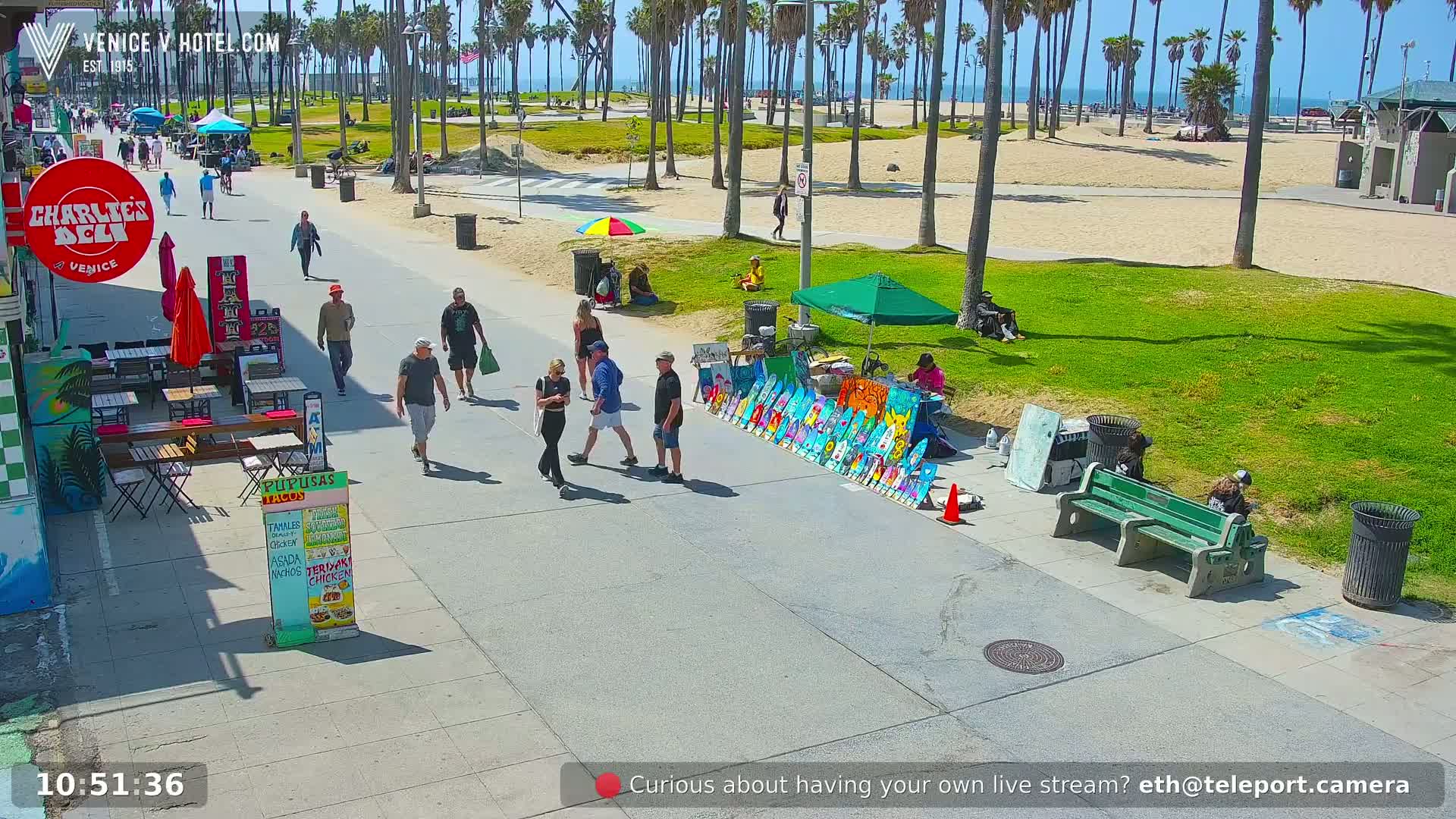 A sunny boardwalk scene shows people strolling near the beach, vendors selling goods, and palm trees lining a grassy area.