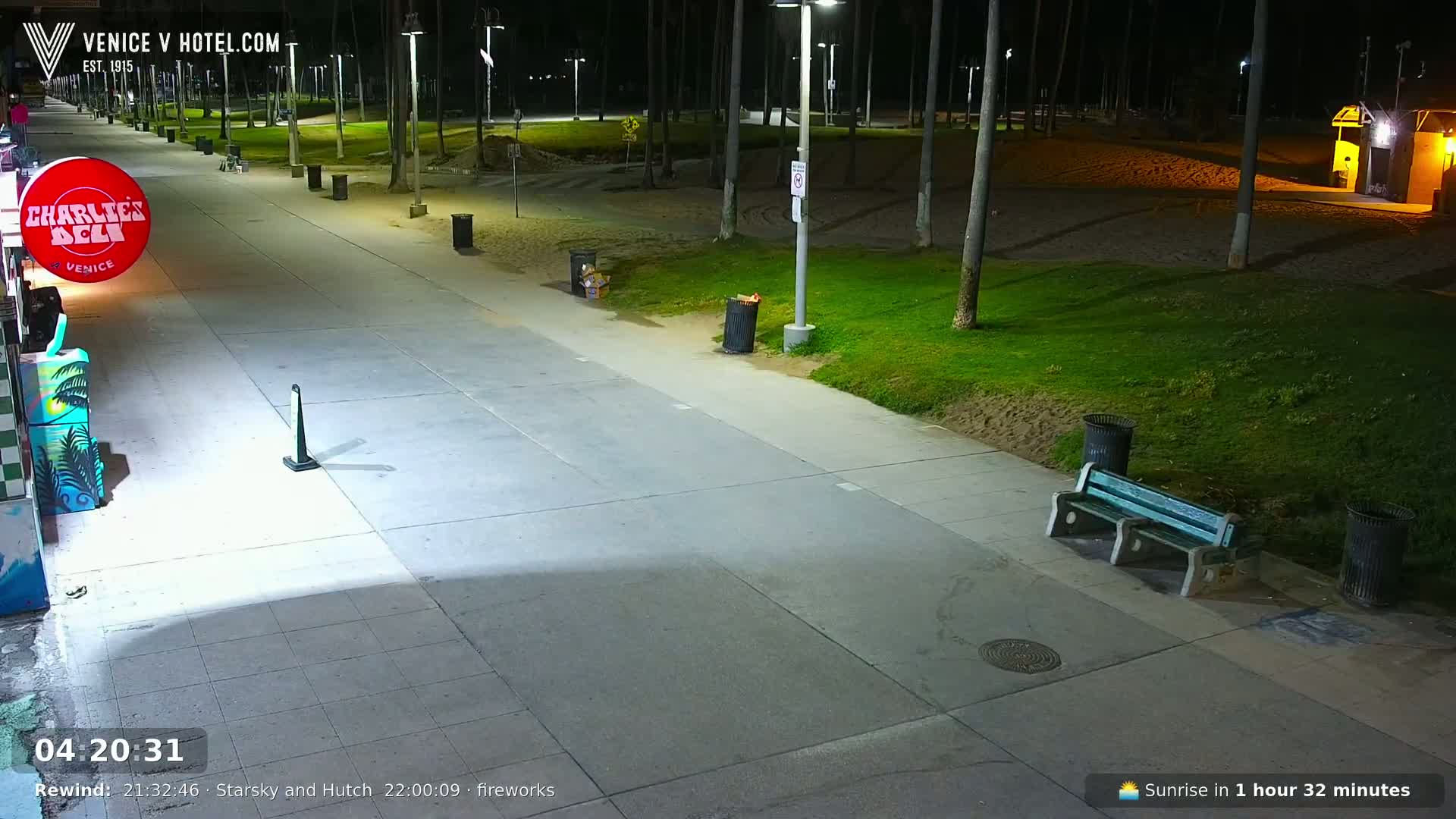A nighttime view of a deserted boardwalk, lined with palm trees and trash cans, next to a grassy area with a bench.
